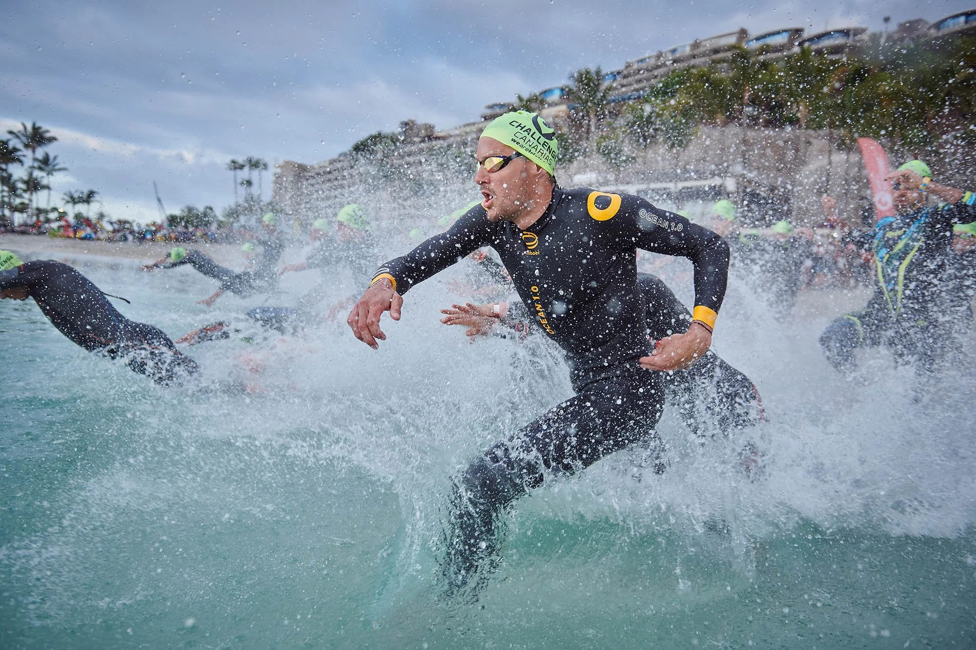 Jan Frodeno entering the water during a race, wearing a black wetsuit, green swim cap, and goggles, with a crowd and buildings in the background.