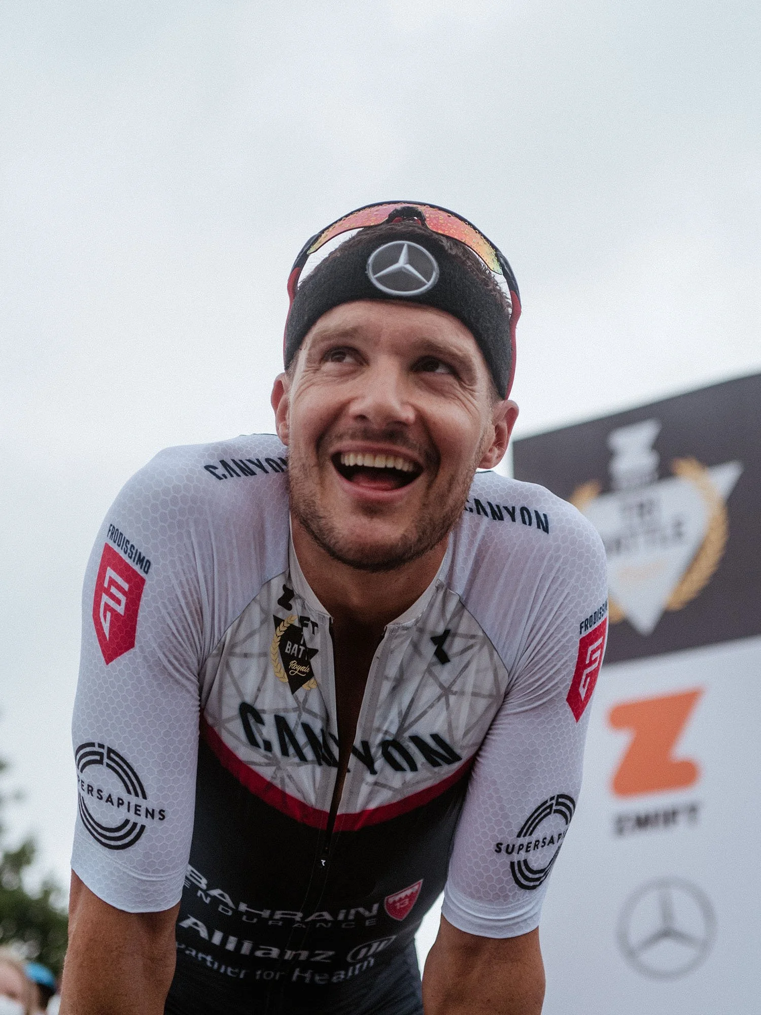 Jan Frodeno smiling and leaning forward, wearing a white race suits with sponsor logos, a black headband with a Mercedes logo, and sunglasses on his head. In the background, there is a blurred banner with logos and a clear sky.