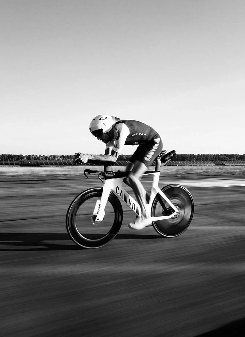 Black and white image of a Jan Frodeno in an aero Oakley helmet and Ryzon racing suit riding a Canyon bicycle along a flat, open landscape.