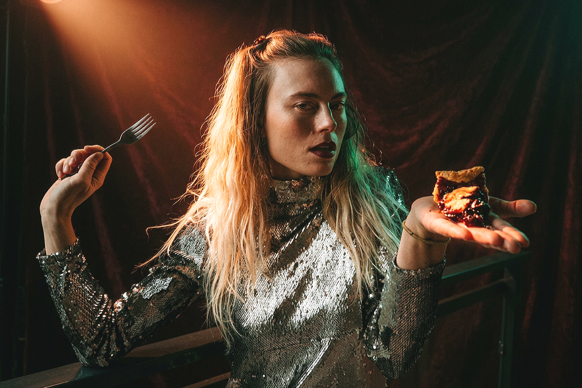 A young woman with wavy blonde hair wearing a shiny, silver, sequin dress holding a plate with a piece of pie and a fork in her right hand, standing in front of a dark, velvet curtain with warm lighting.