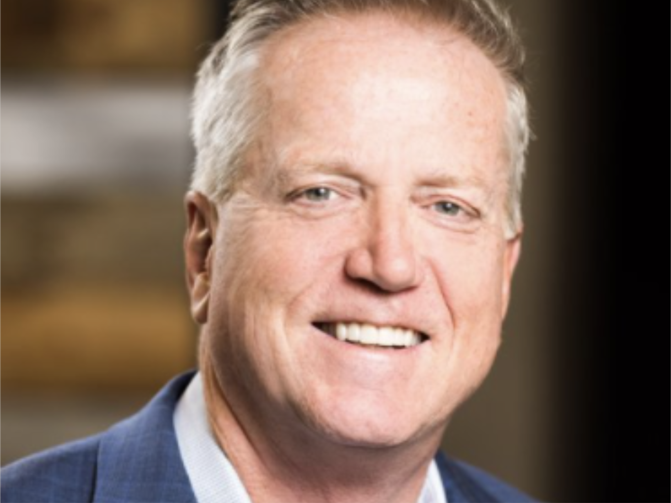 A smiling middle-aged man in a blue suit and light blue shirt, with short, light-colored hair and fair skin, inside a warmly lit room.