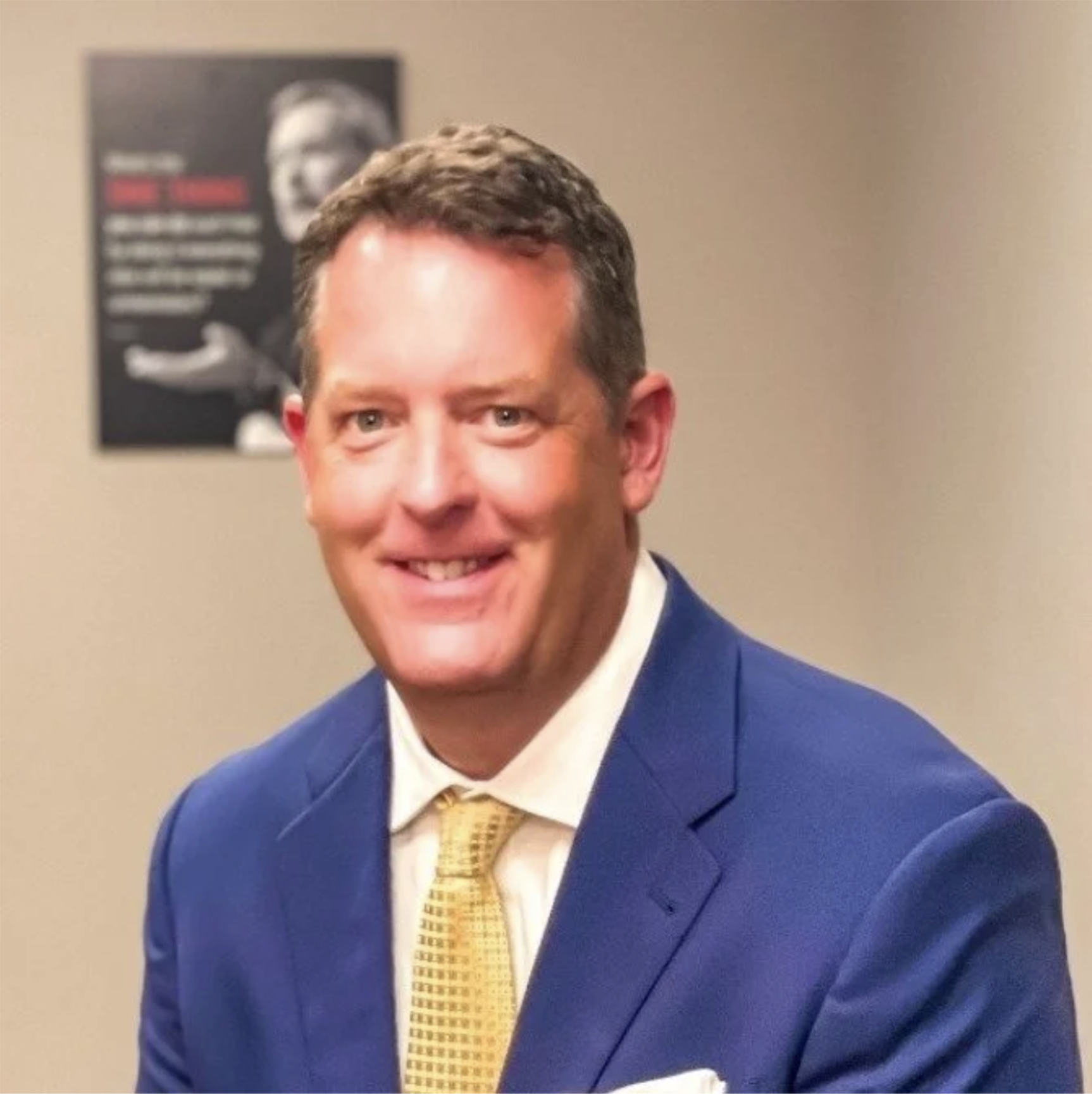 A smiling man in a blue suit with a white shirt and yellow tie, sitting in an office or conference room with a poster on the wall in the background.