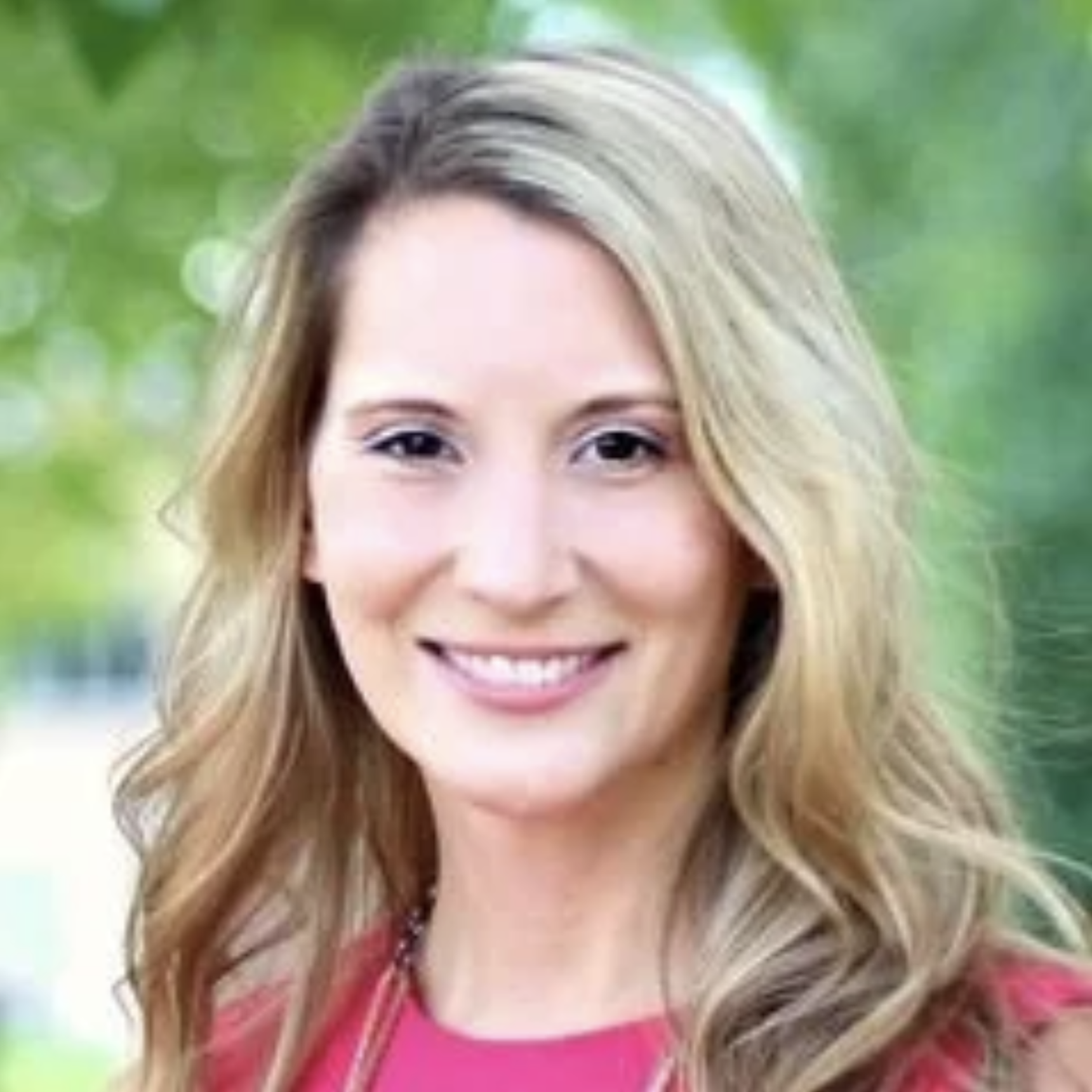 A smiling woman with long wavy blonde hair, wearing a pink top, outdoors with green trees in the background.