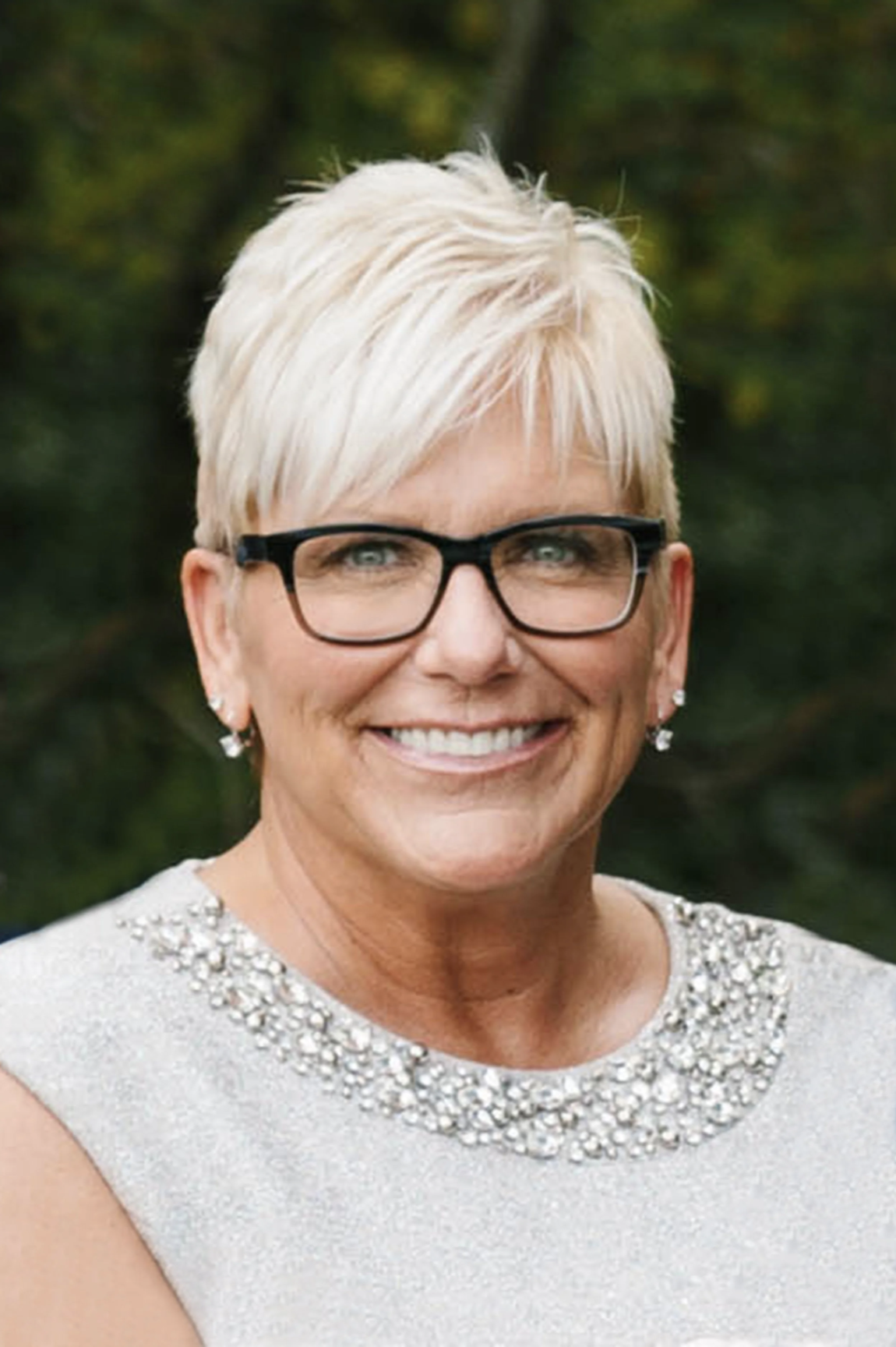 Portrait of a smiling middle-aged woman with short blonde hair, glasses, and earrings, wearing a sparkly dress with a beaded neckline, outdoors with a blurred natural background.