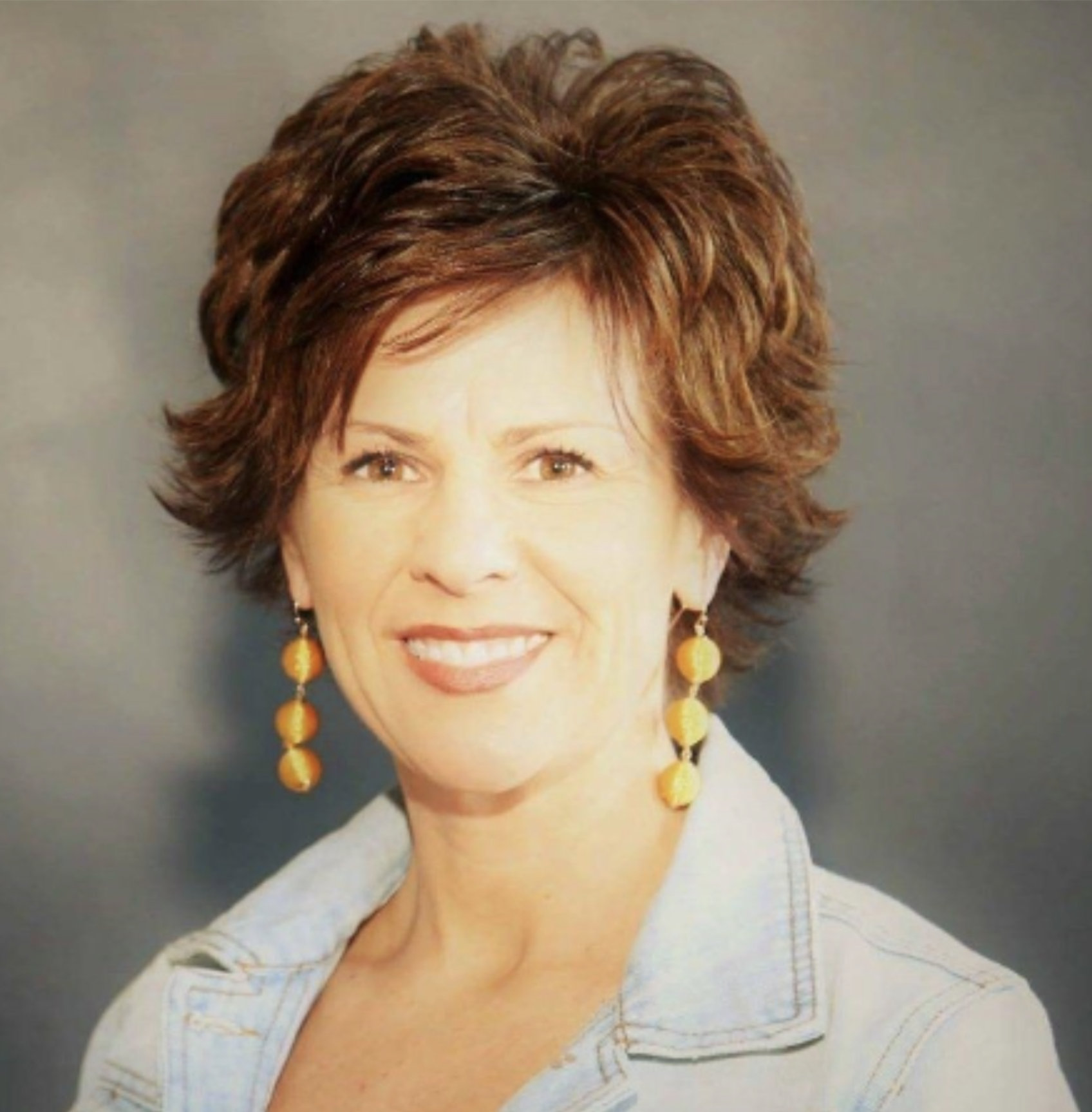 A woman with short, layered, reddish-brown hair smiling, wearing yellow earrings and a light denim jacket, standing against a gray background.