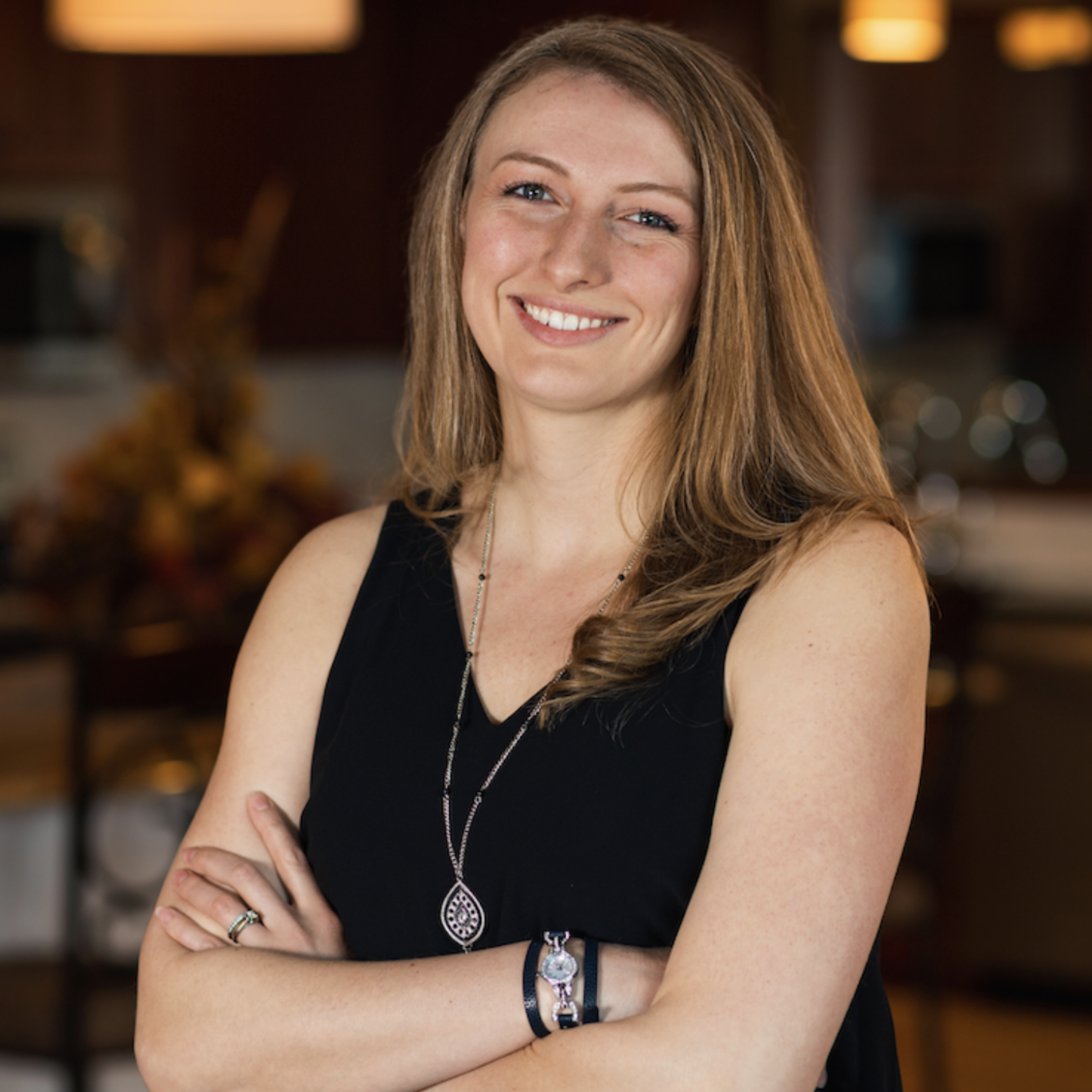 A smiling woman with long, wavy red hair posing with arms crossed in a cozy indoor setting.