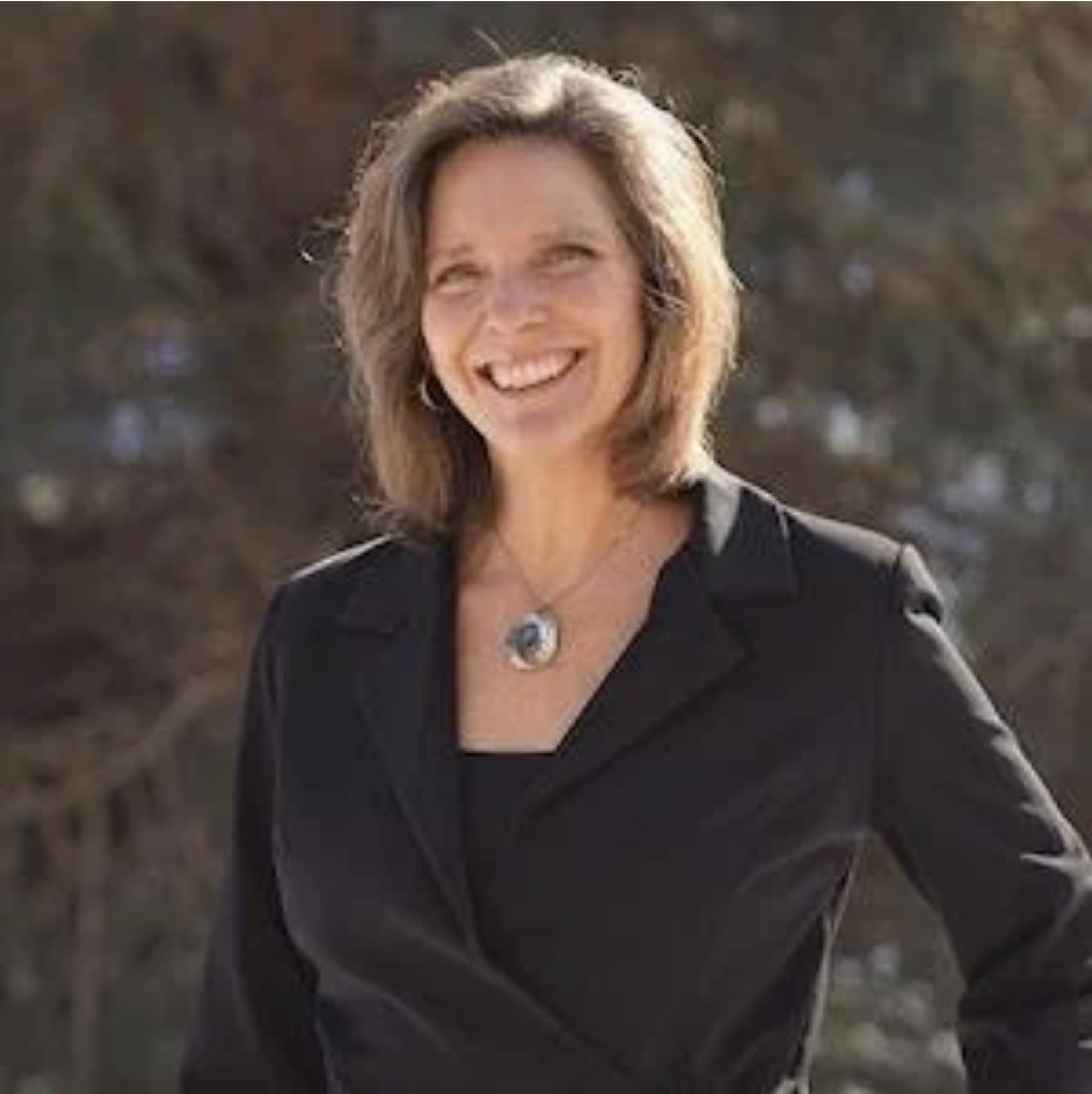 A smiling woman with shoulder-length brown hair, wearing a black jacket and a necklace, outdoors.