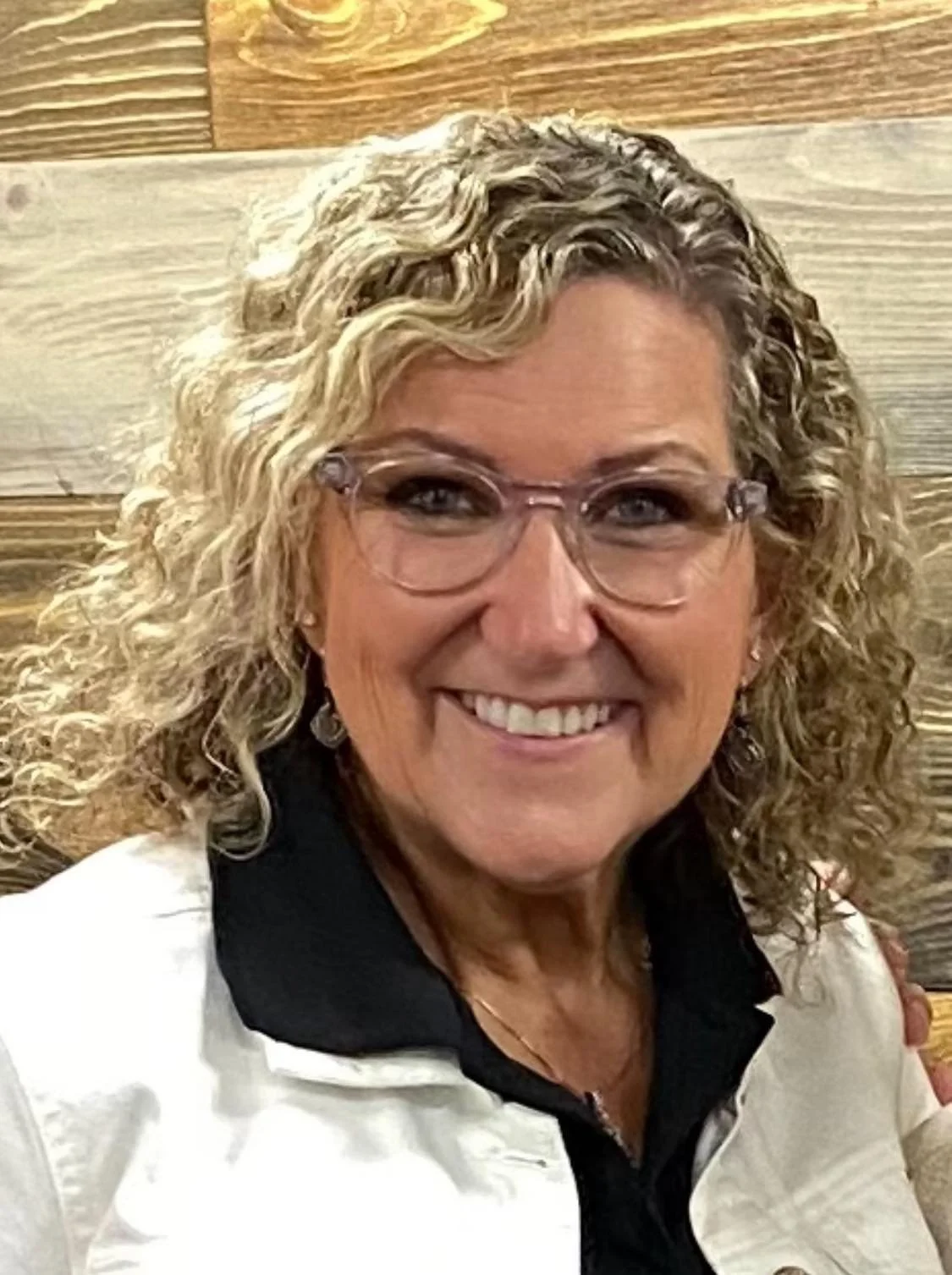 A smiling woman with blonde hair, glasses, and jewelry, wearing a black polka-dot top and a name badge.