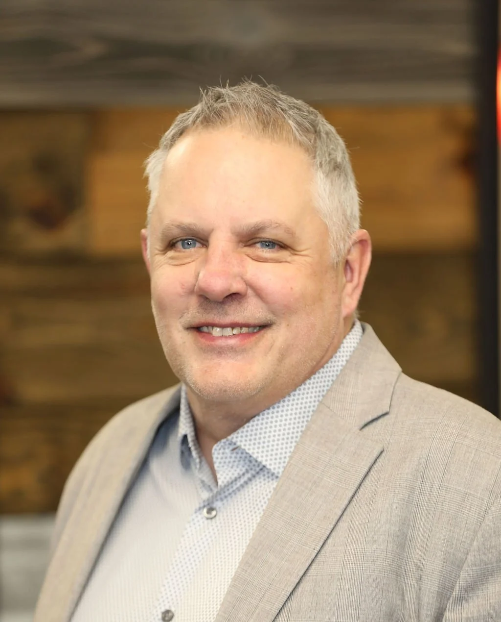 A smiling middle-aged man with short blond hair and blue eyes, wearing a light-colored blazer and a white dress shirt, standing in front of a wooden background.