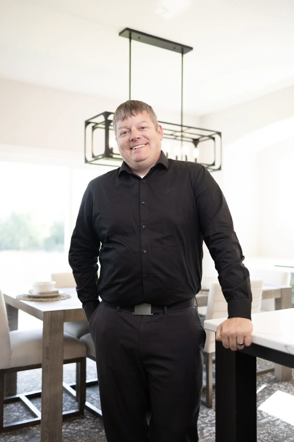 Portrait of a man in a black shirt and black pants standing in a bright dining room with modern light fixture and a wooden dining table with white dishes.