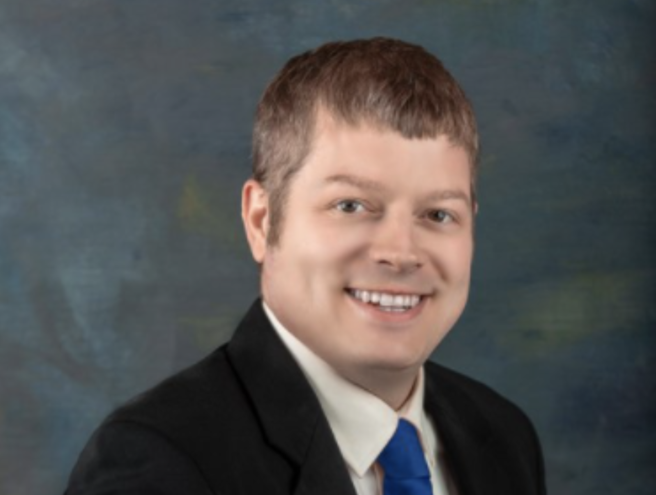 Professional portrait of a young man with short brown hair, wearing a black suit, white shirt, and blue tie, smiling against a dark green and blue painted background.