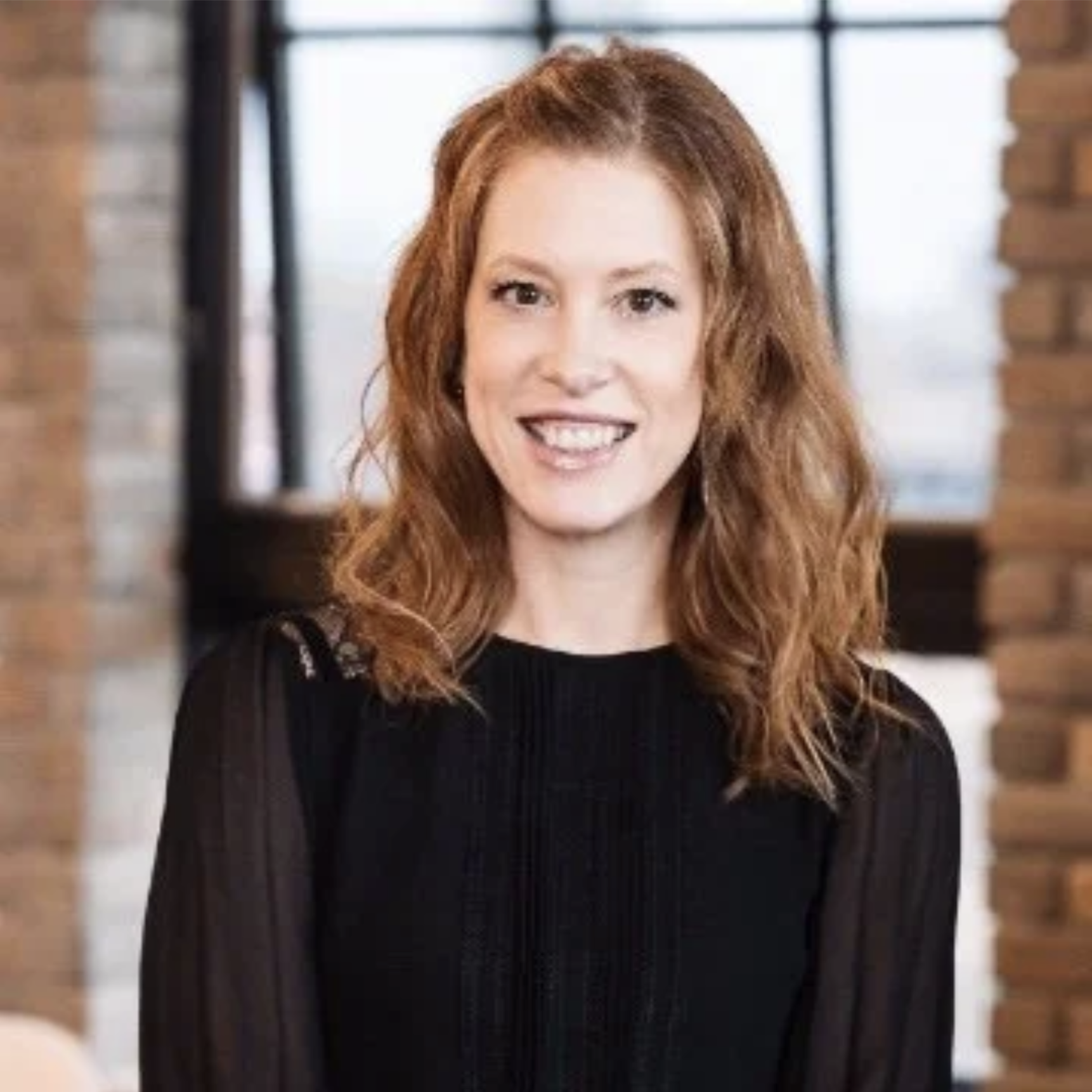 Red-haired woman smiling in a black top, standing indoors with brick and large windows in the background.