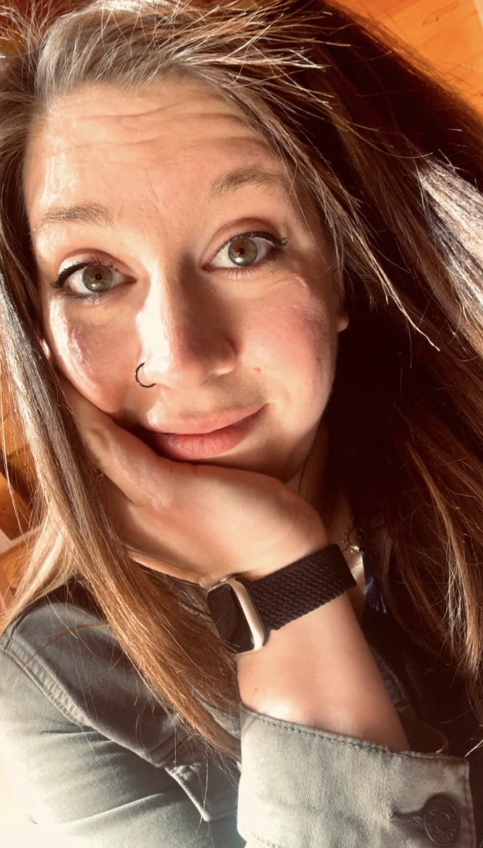 A woman with light brown hair and fair skin, resting her chin on her hand, wearing a black wristband, a nose ring, and a necklace, with a wooden surface in the background.