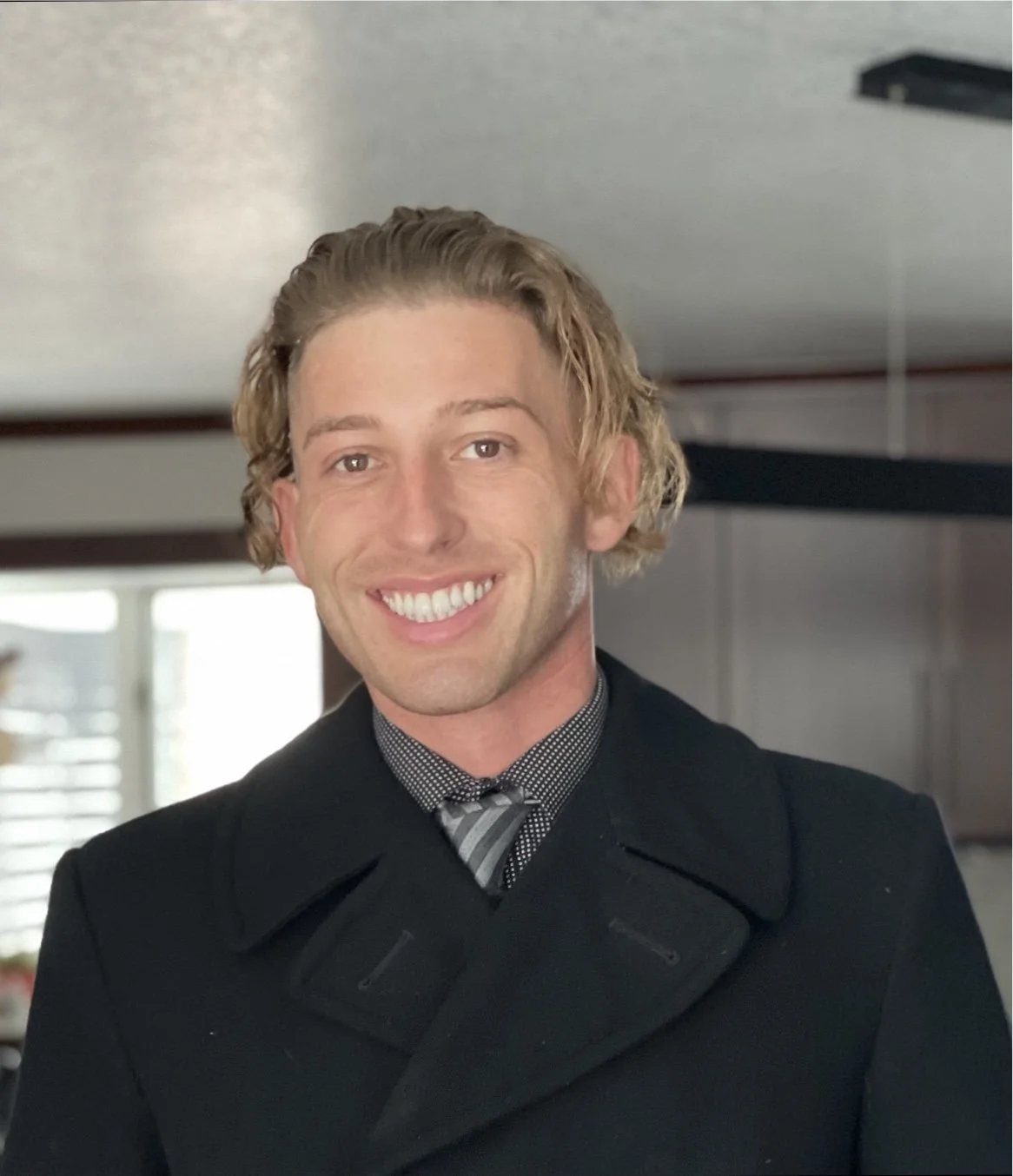 A young man with wavy blonde hair and a big smile, wearing a black coat and a polka dot shirt with a striped tie, standing indoors.