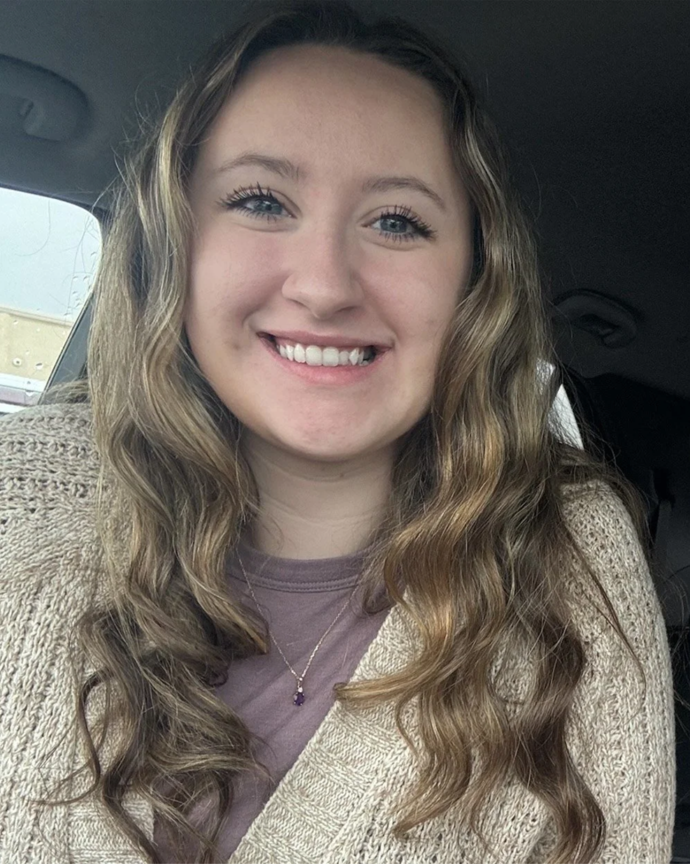 A young woman with wavy, light brown hair and blue eyes smiling inside a vehicle.