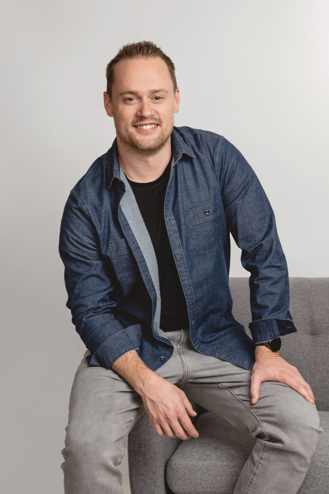 A man sitting on a gray couch against a plain light-colored wall, wearing a dark denim shirt over a black t-shirt, and light gray pants.