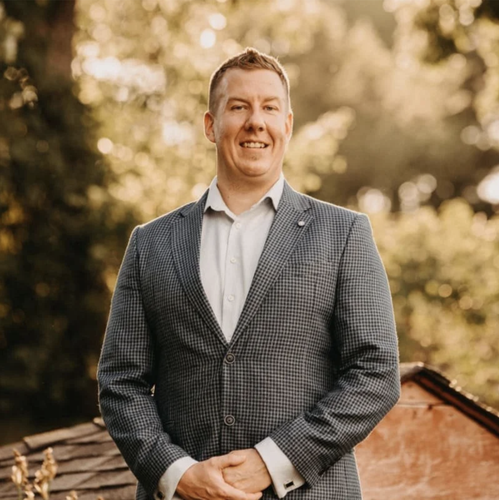 A smiling man wearing a checkered blazer and white shirt standing outdoors with blurred trees and sunlight in the background.