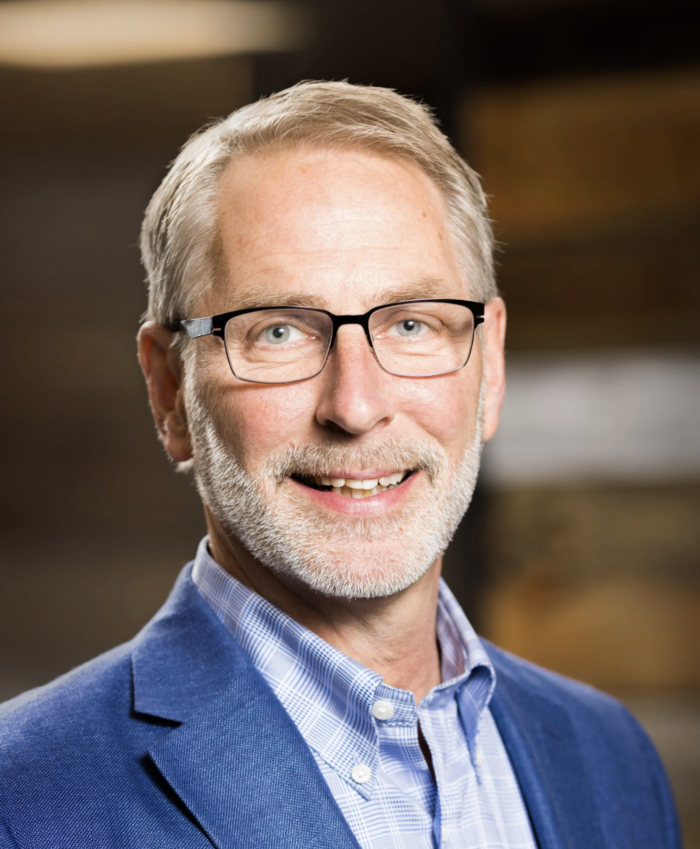 Portrait of a middle-aged man with gray hair, glasses, and a beard, wearing a blue blazer and checked dress shirt, smiling.