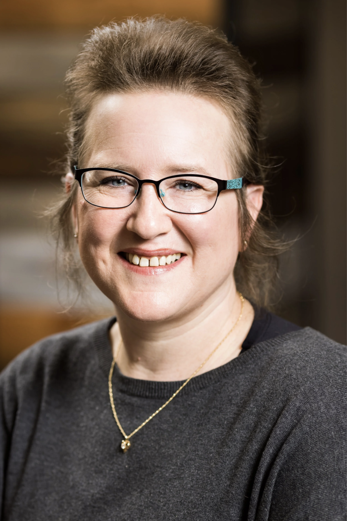 A woman with glasses smiling, wearing a dark sweater and gold necklace, in an indoor setting.