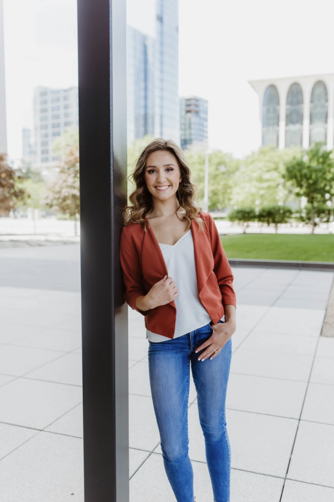 A young woman with wavy brown hair smiling outdoors, leaning against a black vertical pole, with city buildings and green trees in the background.