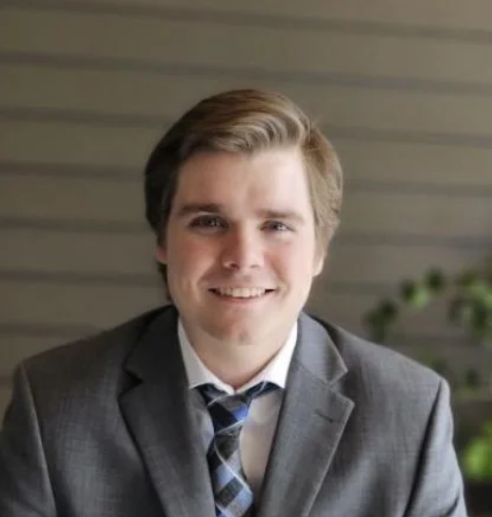 A young man with light brown hair, dressed in a gray suit, white shirt, and tie, smiling in an indoor setting.