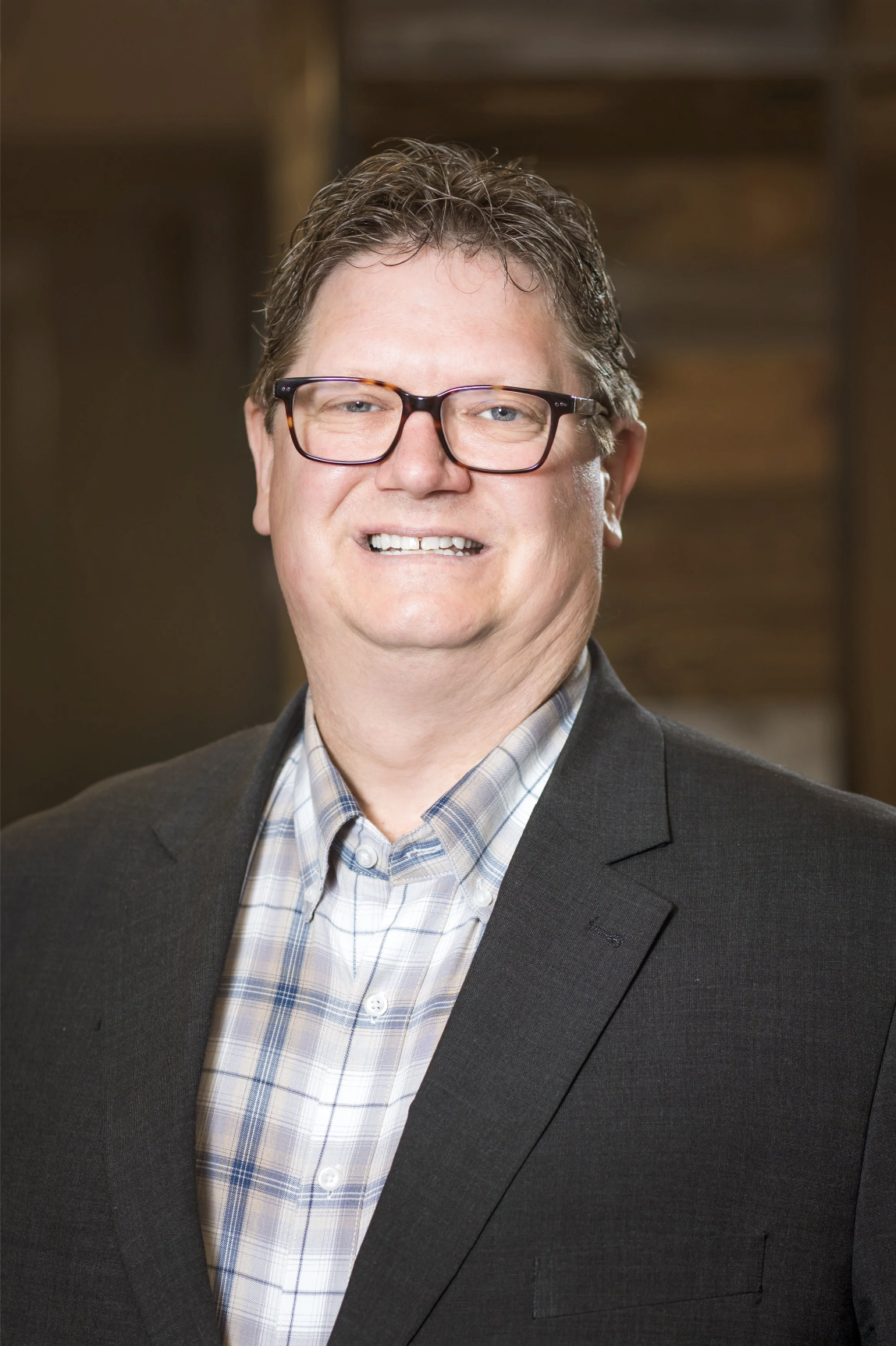 A middle-aged man with glasses, smiling, in a suit and plaid shirt, posing indoors with a blurred background.