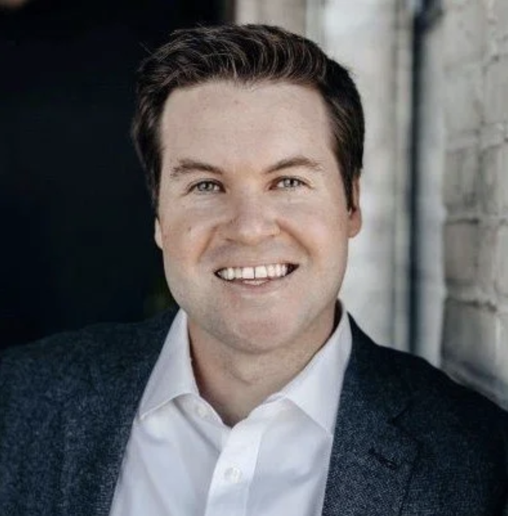 Headshot of a man with short dark hair wearing a white shirt and dark blazer, smiling against a blurred outdoor background.
