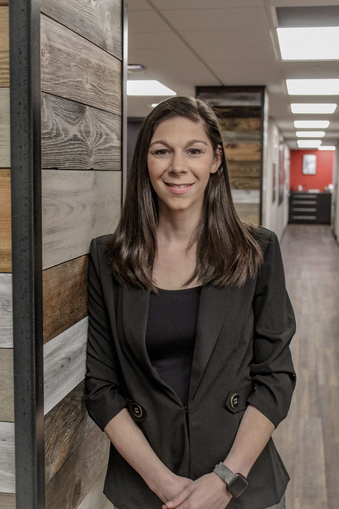 A woman with long dark hair wearing a black blazer and black shirt standing in an office hallway, leaning against a wooden wall, smiling at the camera.