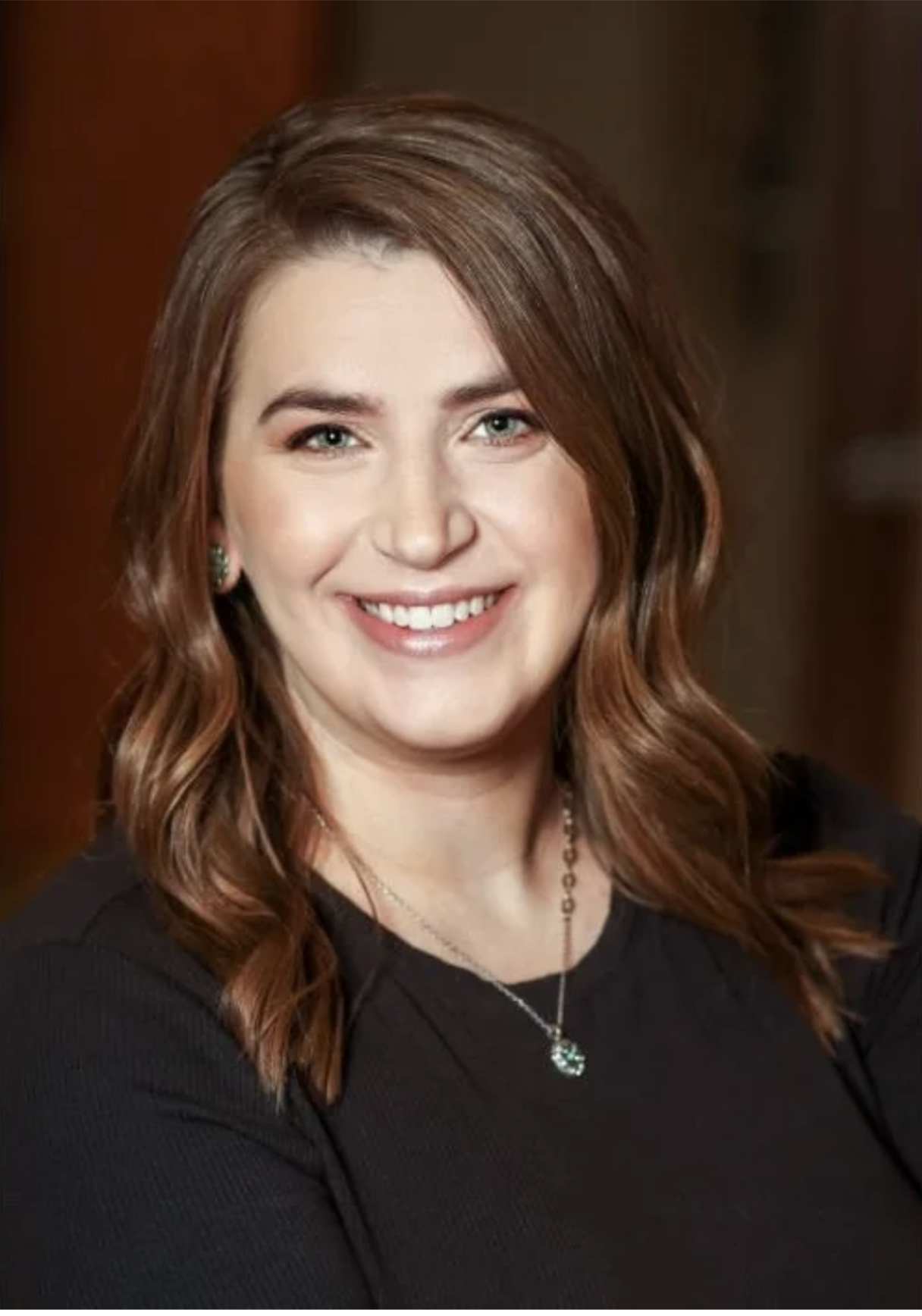 A woman with shoulder-length reddish-brown hair, green eyes, and a bright smile, wearing a black top and jewelry, in an indoor setting.