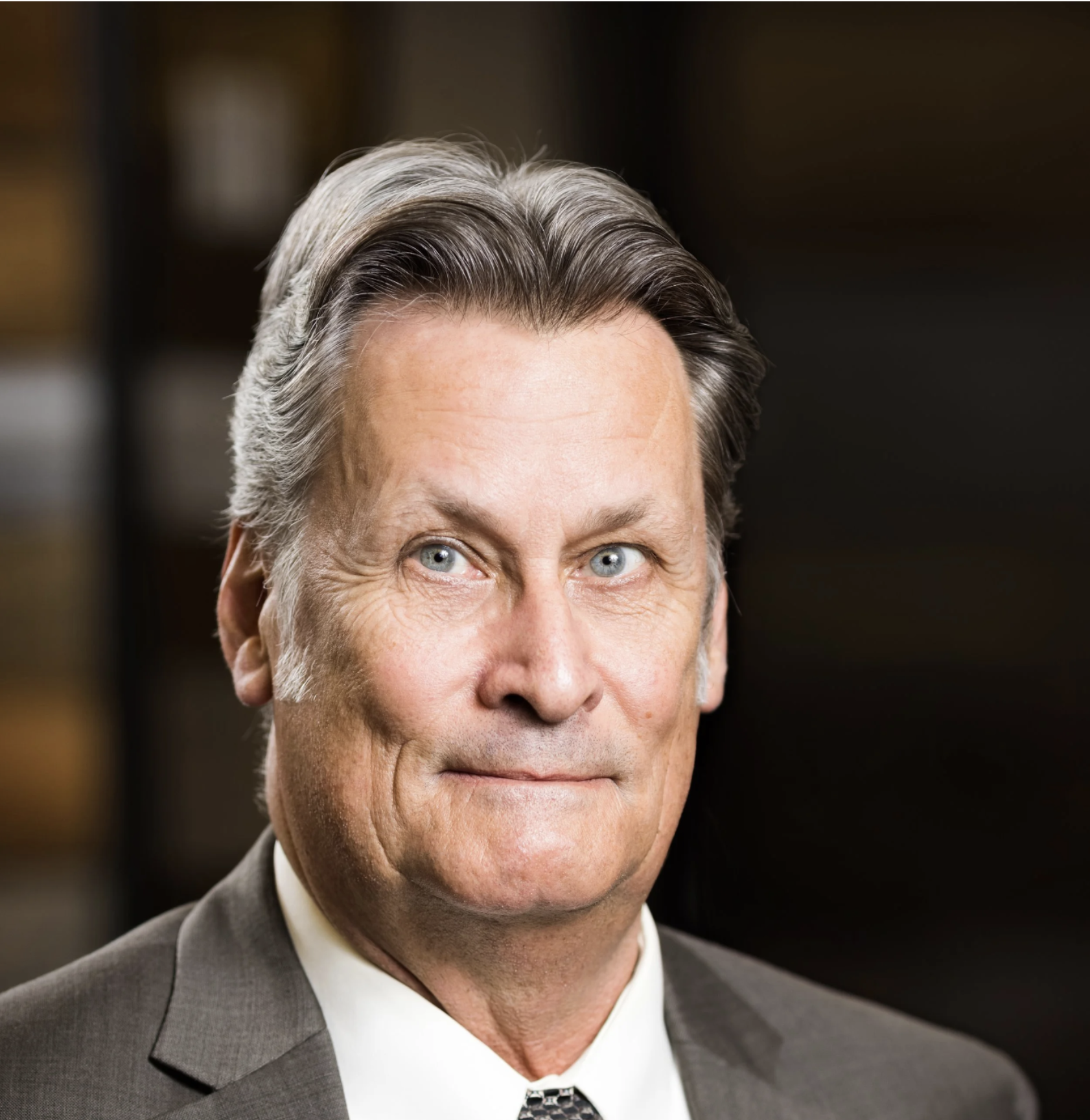 Close-up of a middle-aged man with gray hair and blue eyes, wearing a gray suit and tie, smiling slightly in a professional setting.