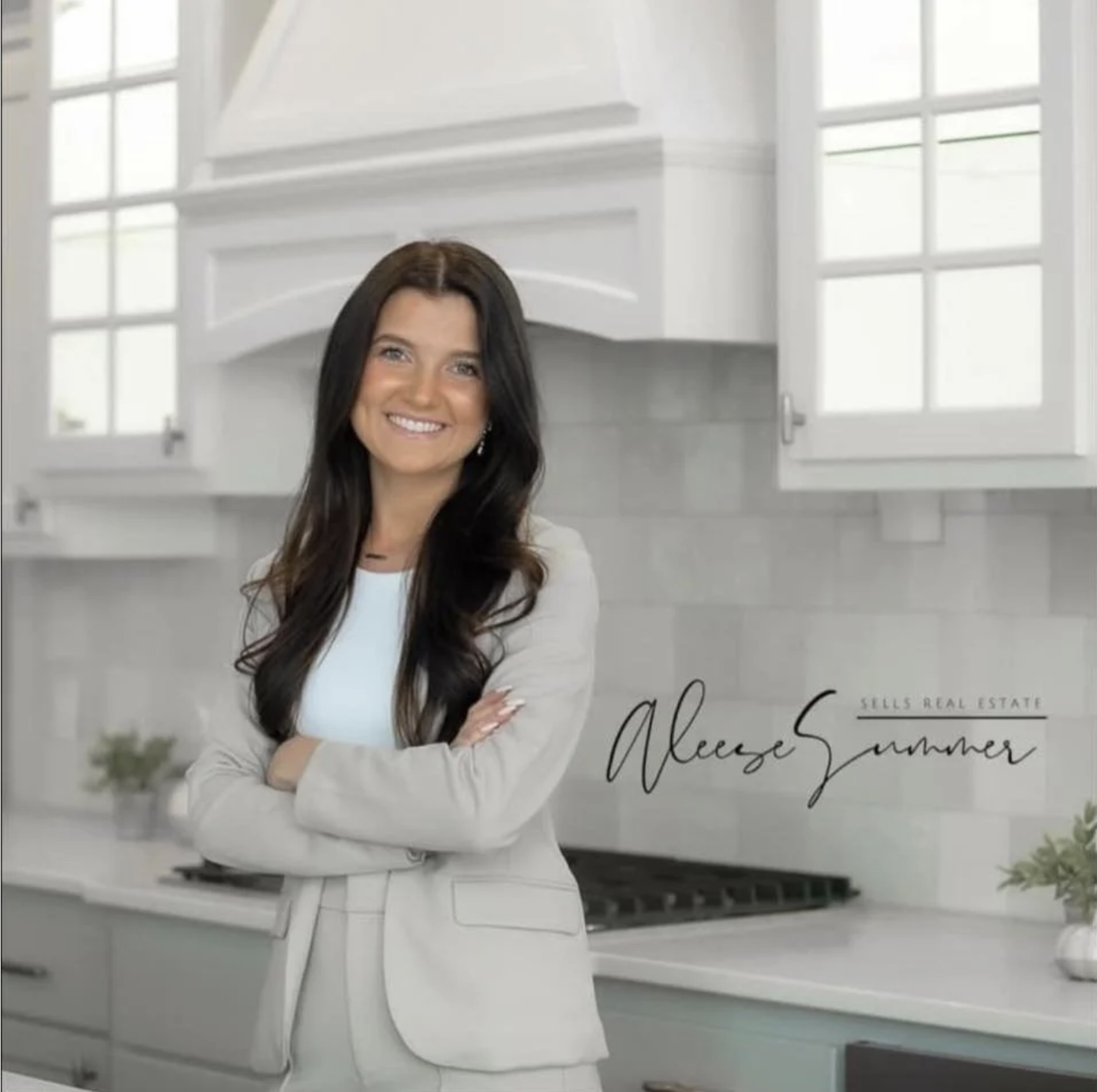 A woman with long dark hair, wearing a light-colored blazer and blouse, stands in a modern white kitchen with crossed arms, smiling at the camera.