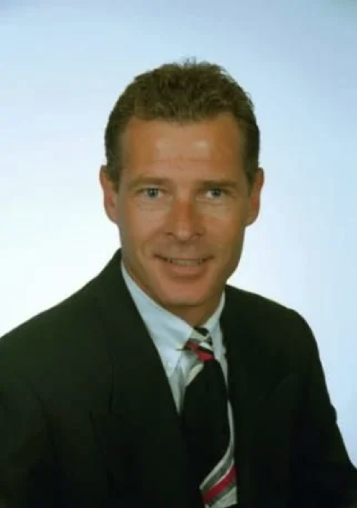 A man with short, dark hair wearing a black suit, white shirt, and striped tie, smiling against a light blue background.
