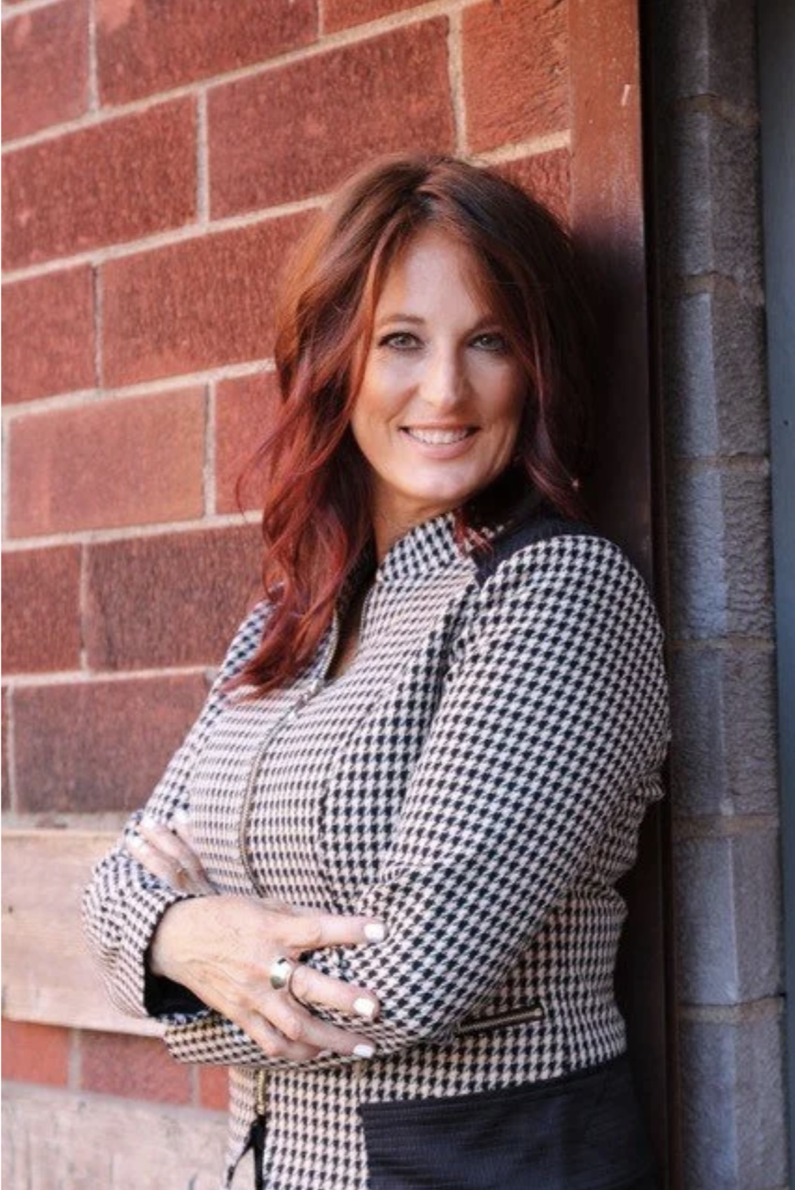 A woman with shoulder-length red hair, wearing a black and white houndstooth patterned jacket, standing with arms crossed and leaning against a brick wall, smiling at the camera.