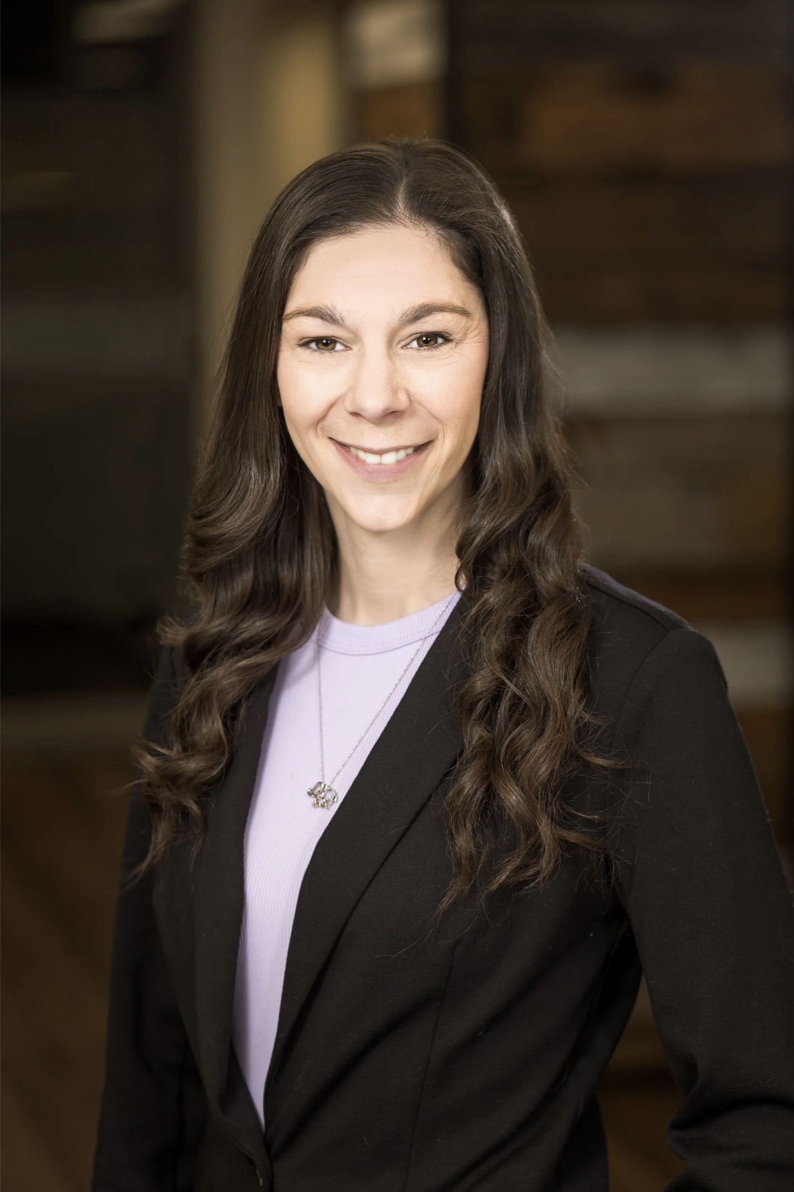 A woman with long, wavy brown hair smiling at the camera, wearing a black blazer, light purple shirt, and a necklace with a small pendant, standing indoors with a blurred background.