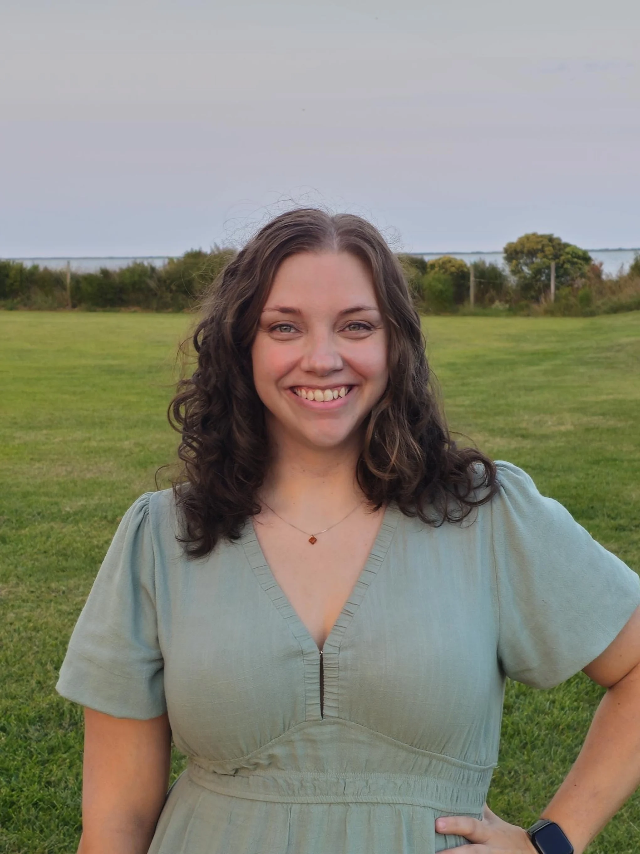 Smiling woman with curly brown hair in a light green dress standing on a grassy field outdoors during daytime.