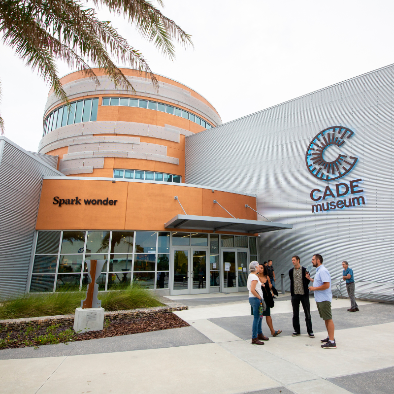 The CADE Museum's entrance with a group of people talking outside, modern architecture, palm tree, and a sculpture nearby.