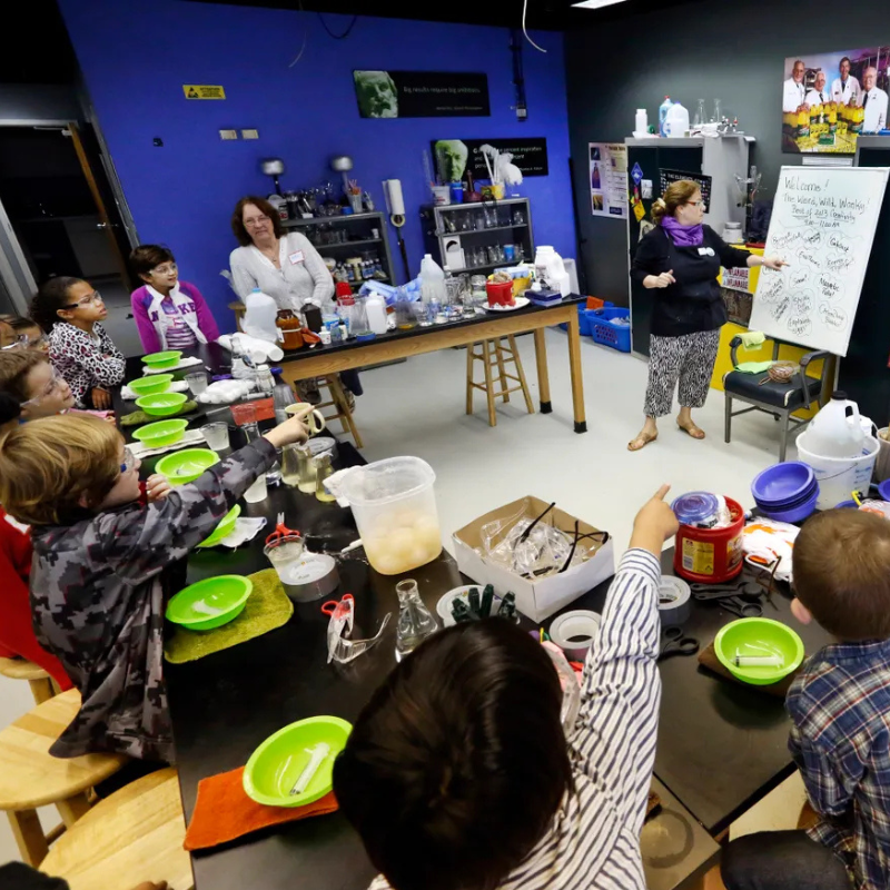 Children participating in a STEAM camp at the Cade Museum, with a teacher pointing at a whiteboard displaying notes, in a brightly lit classroom with science supplies and equipment.