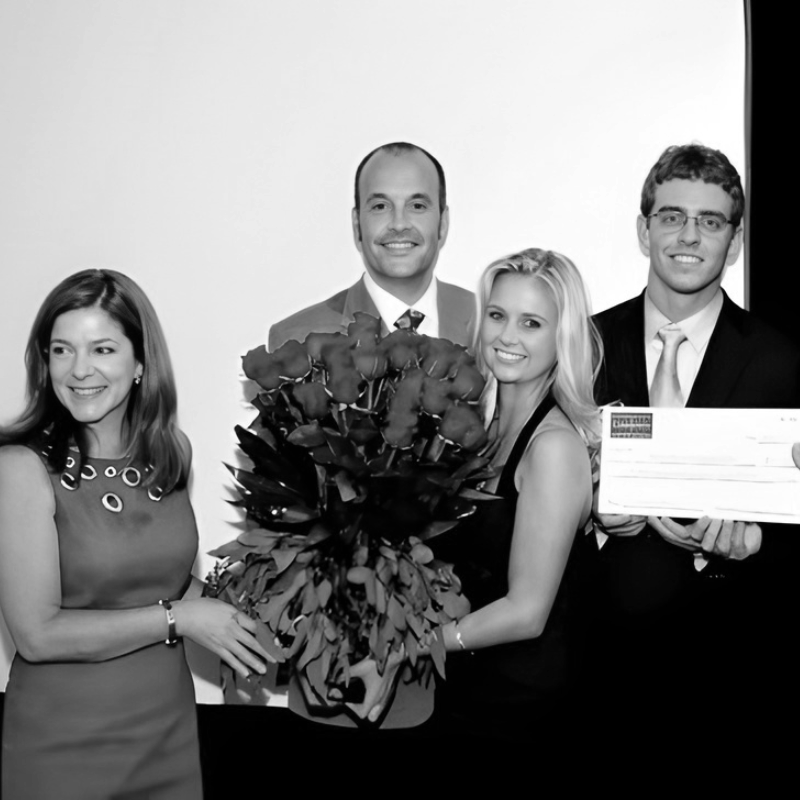 Group of four people, two women and two men, celebrating with a large bouquet of roses and a check, smiling at the first Cade Prize competition in 2010.
