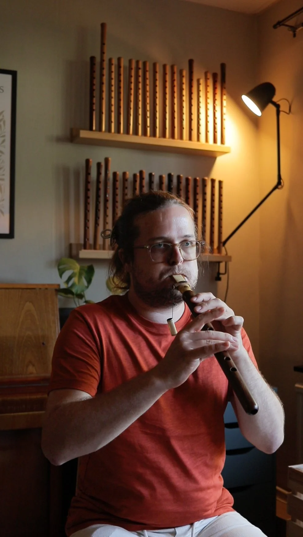 A man playing a woodwind instrument, sitting indoors with acoustic panels on the wall behind him, a floor lamp to his right, and a small plant nearby.