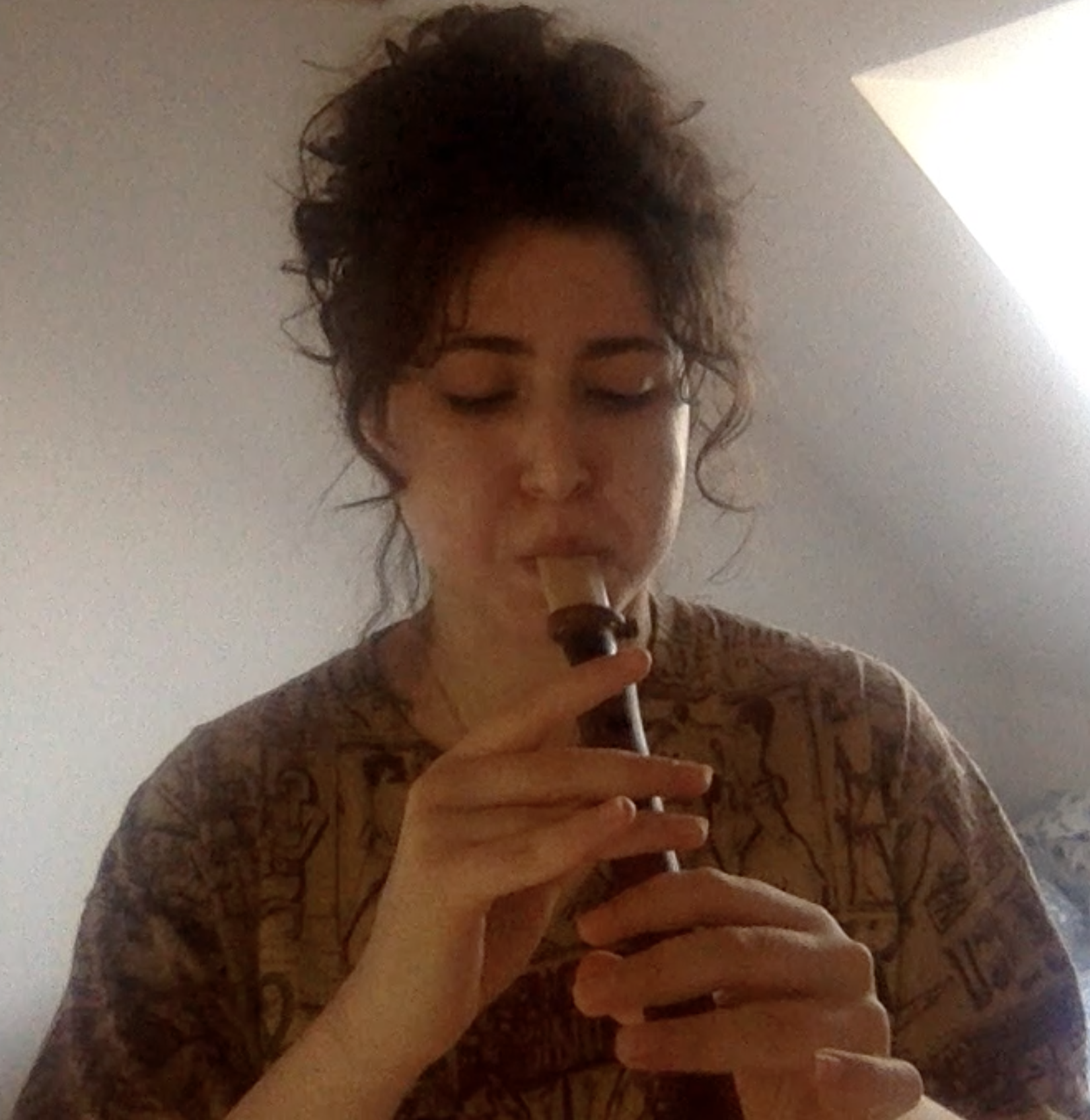 A young woman with curly hair playing a recorder, with her eyes closed, in an indoor setting.