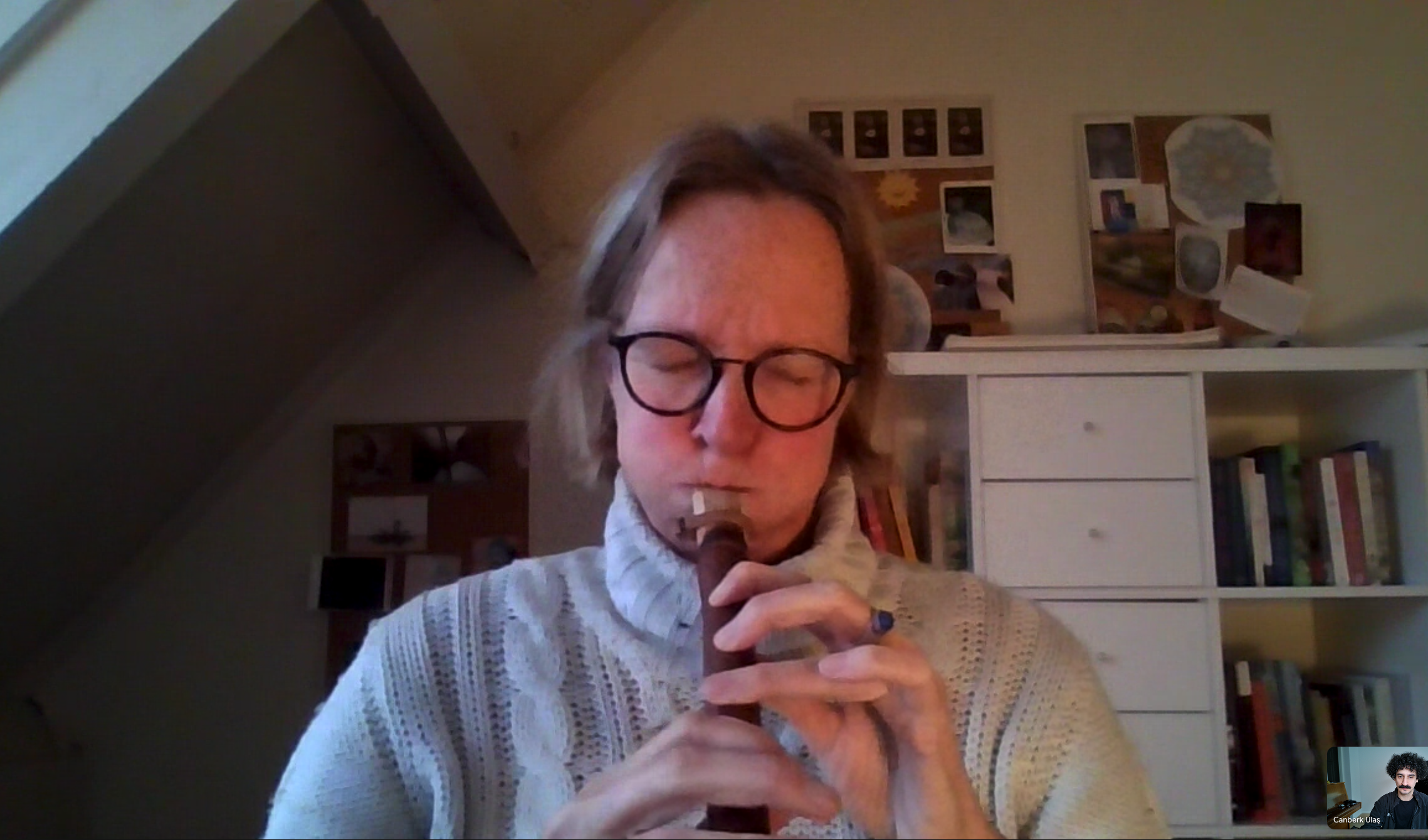 A woman with glasses playing a wooden wind instrument, possibly a recorder, in a cozy room with bookshelves and art on the wall.