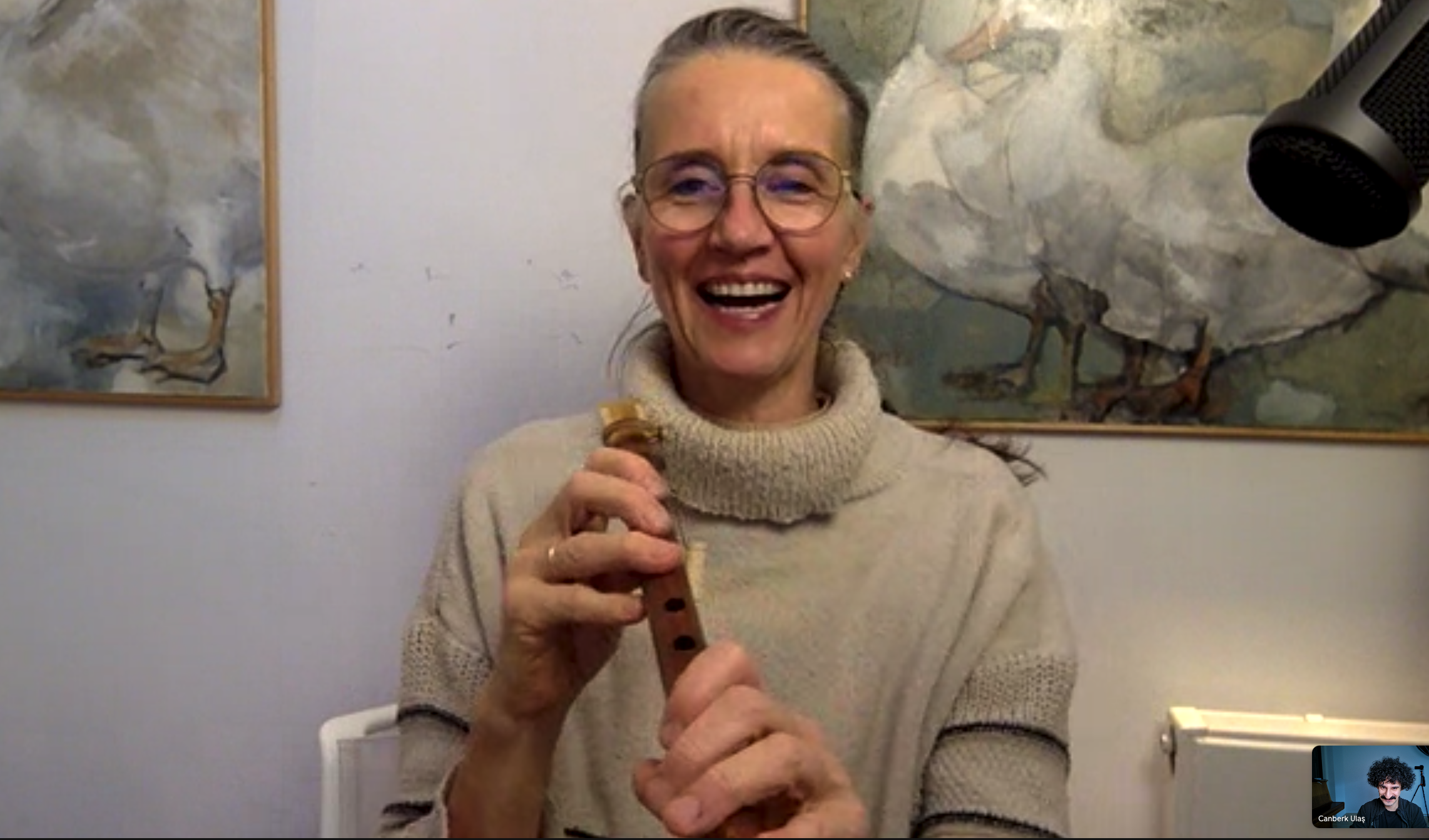 A smiling woman wearing glasses and a beige turtleneck sweater playing a recorder, with abstract paintings on the wall and a black microphone in the background.