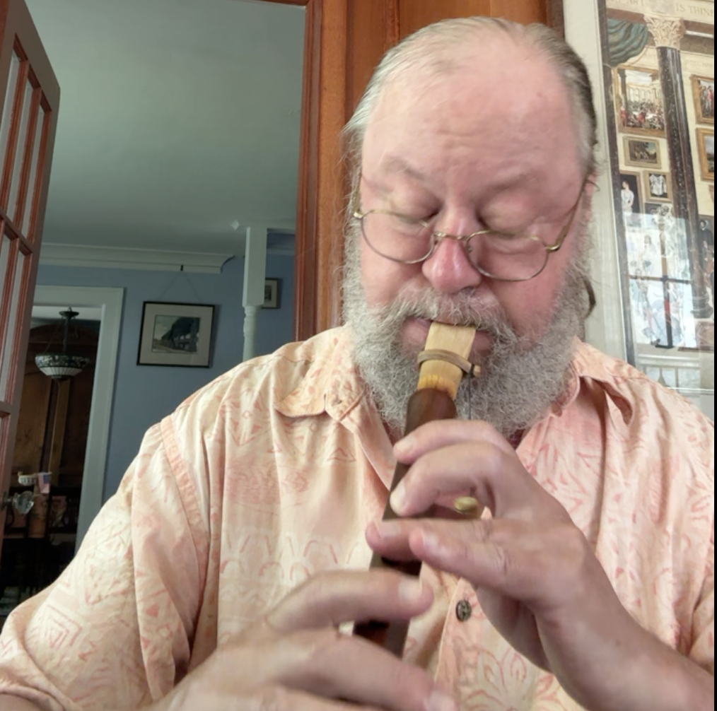 An older man with white hair, glasses, and a beard is playing a wooden recorder.