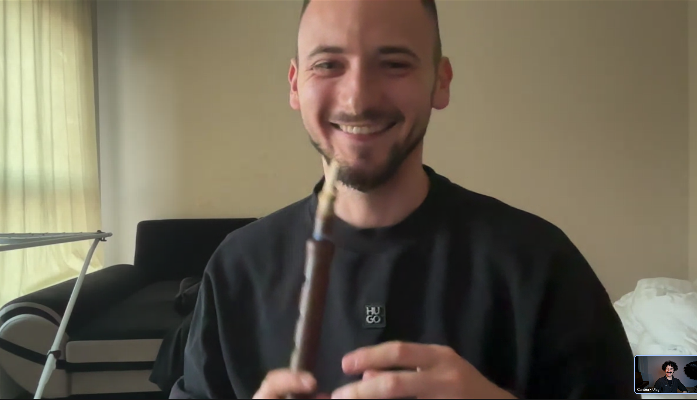 A man smiling while holding a smoking electronic cigarette or vape pen, sitting in a room with beige walls, a black couch, and a window with yellow blinds.
