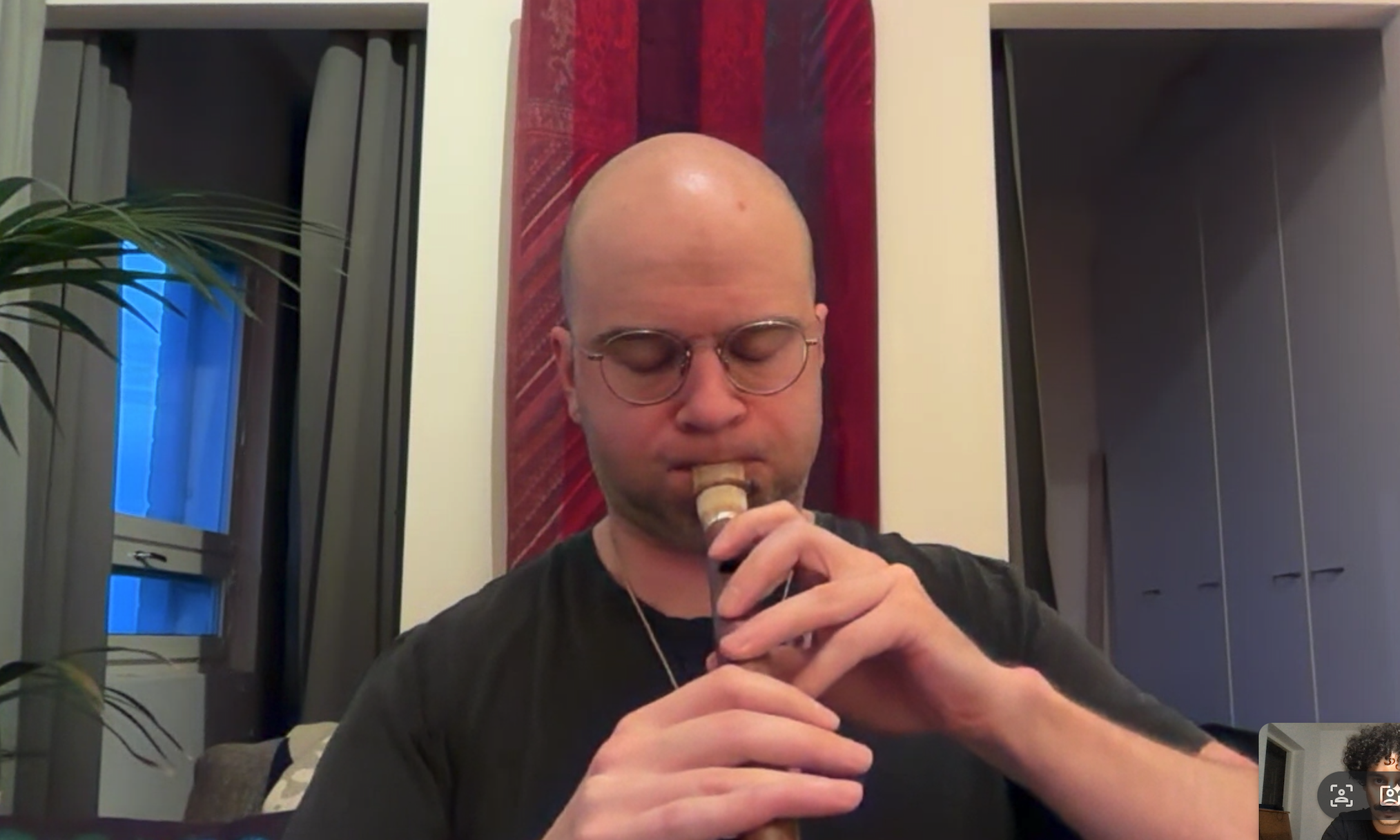 A bald man with glasses is playing a recorder in a room with a window, plants, and a red wall hanging.