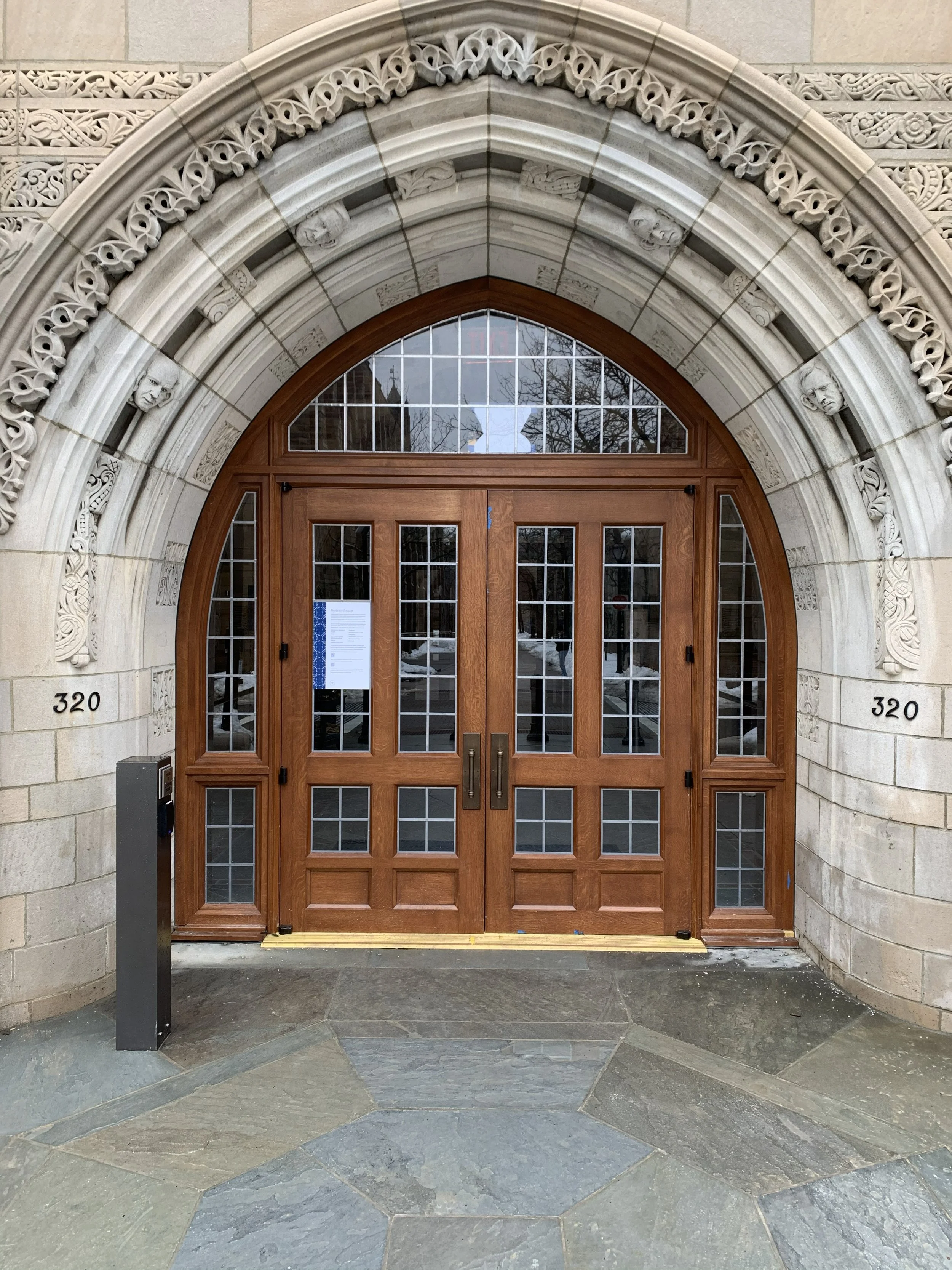 The entrance to a building with a large wooden door set within a decorative stone archway. The arch has intricate carvings and faces along its span. The door has glass panels and is framed by a stone that is marked with the number 320 on both sides.