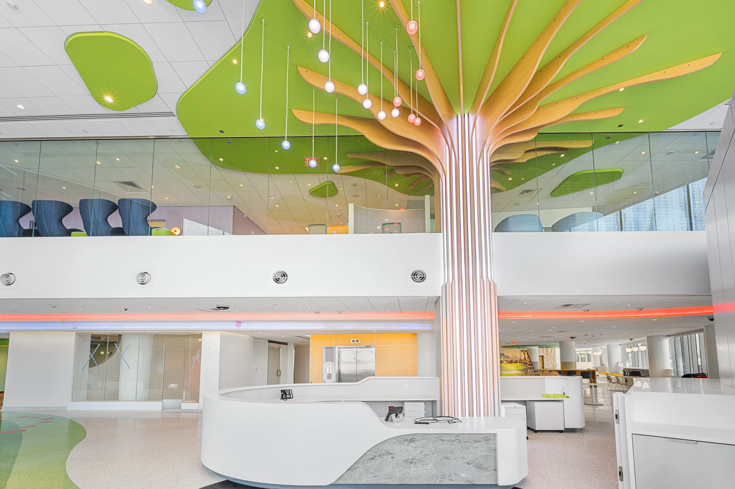 Interior of a modern office building lobby featuring a large decorative tree with a green canopy and wooden branches extending from a striped trunk, illuminated hanging lights, and a reception desk in the foreground.