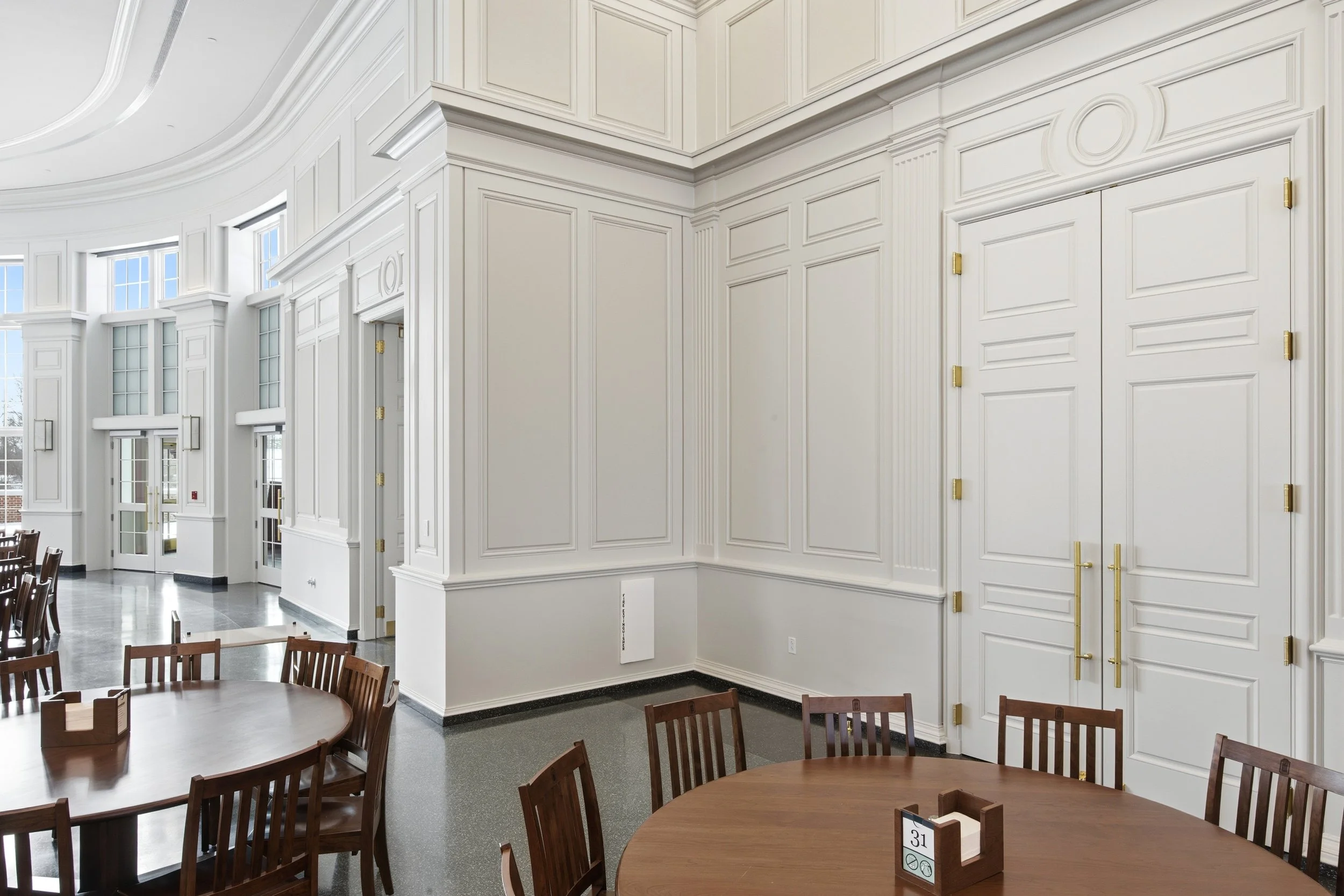 Interior of a formal dining area with large white walls, detailed wall paneling, and wooden chairs around round tables.
