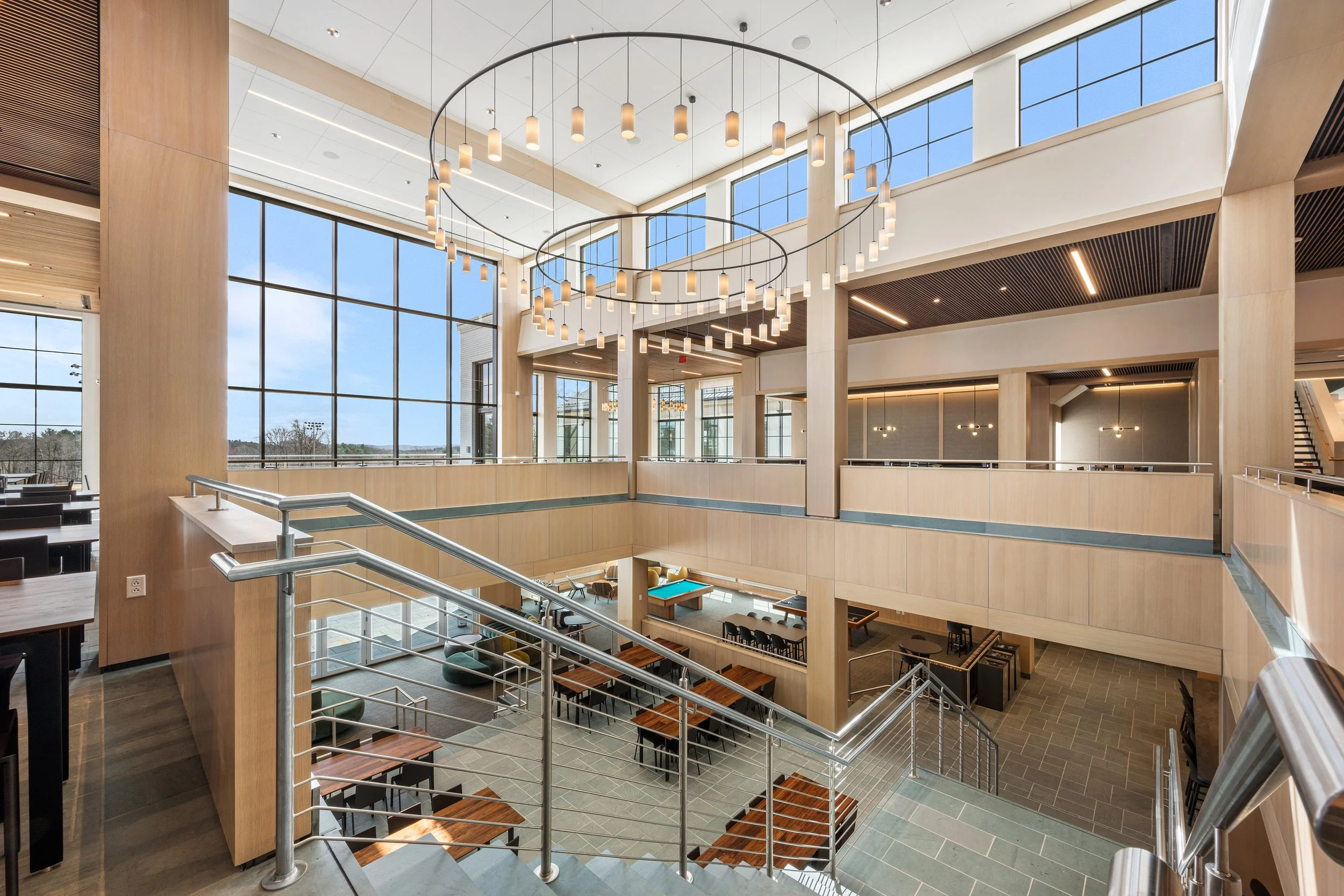 Interior of a modern multi-story building with large windows, wooden accents, and a staircase, featuring tables and chairs, with hanging circular light fixtures.