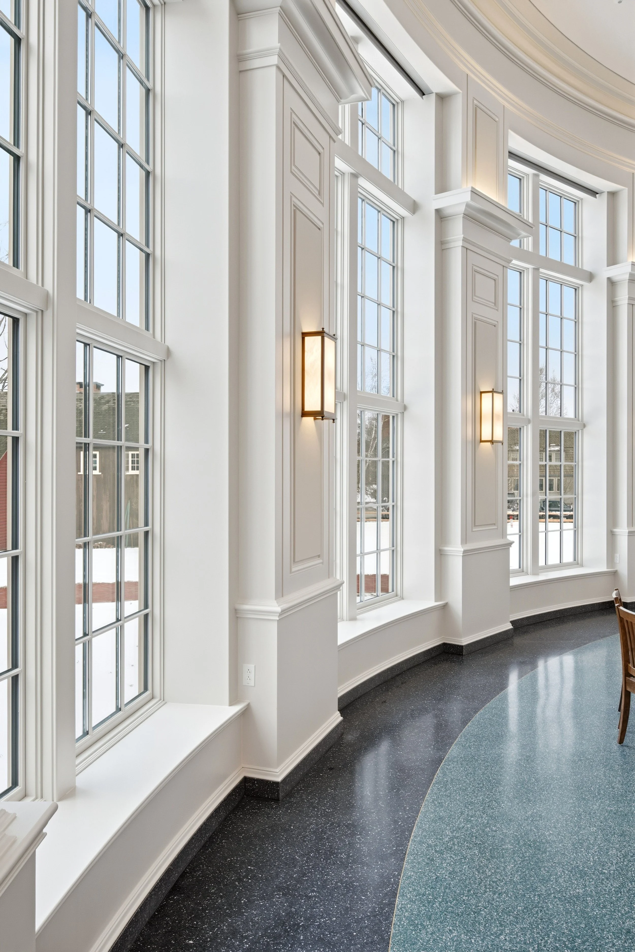 Bright interior with large multi-pane windows, white walls with decorative molding, and modern rectangular wall sconces, with a curved black and teal terrazzo floor.
